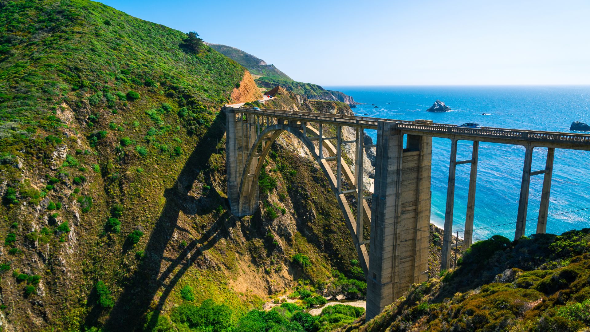 A scenic view of the Bixby Creek Bridge, a reinforced concrete open-spandrel arch bridge, spanning a deep canyon along the California coastline, with the Pacific Ocean visible in the background and lush green hills on either side. 