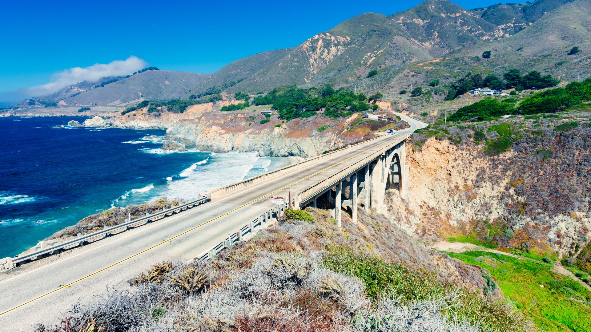 A scenic view of the Rocky Creek Bridge, a concrete arch bridge, spanning a canyon along the Pacific Coast Highway (Highway 1) in Big Sur, California. The deep blue Pacific Ocean is visible on the left, with waves crashing against the rocky coastline, and lush, mountainous terrain rises on the right. The bridge carries a two-lane road, and the sky is clear and bright blue.