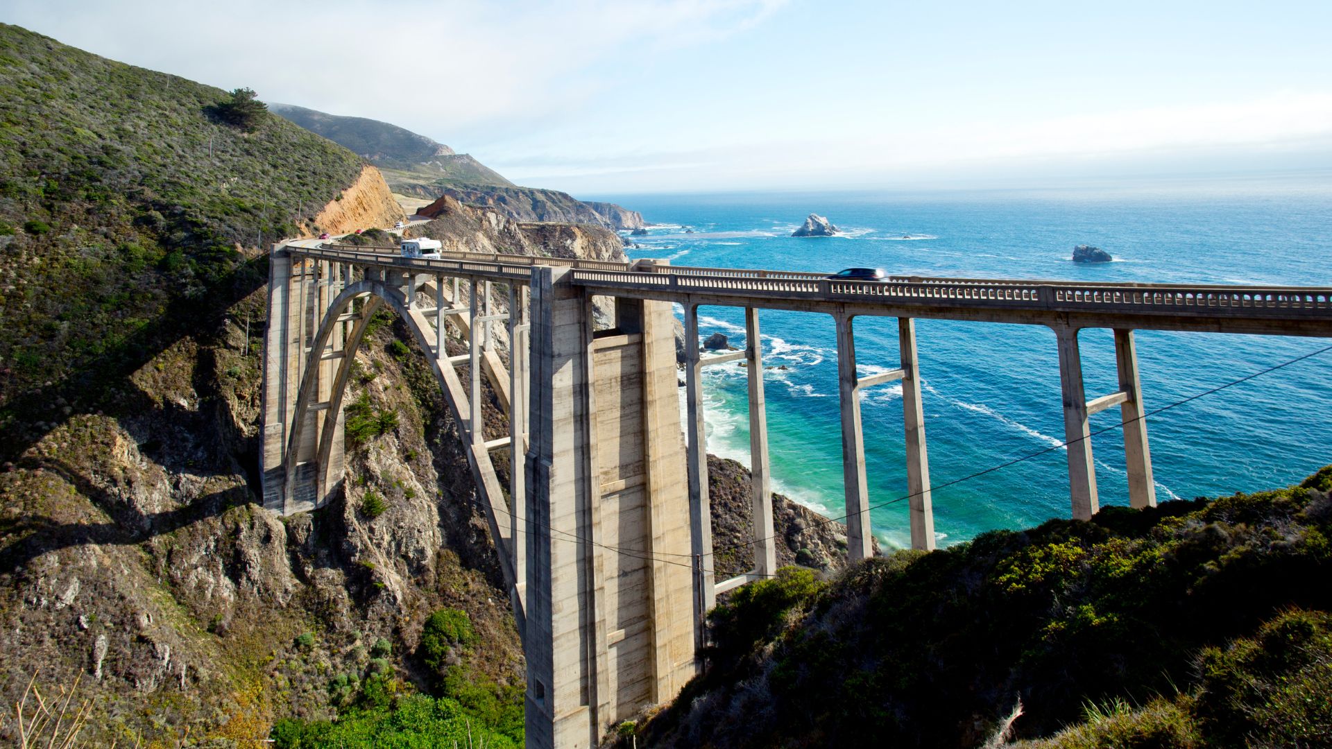 A concrete arch bridge, the Bixby Creek Bridge, spans a deep coastal canyon above the Pacific Ocean, with scenic cliffs and the California coastline visible under a clear sky.