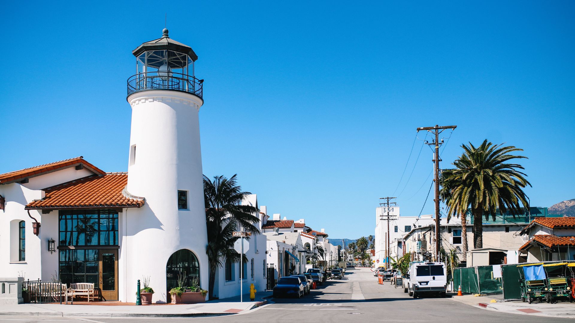 A white lighthouse-style building, which is the Bluewater Grill Seafood Restaurant, stands prominently on a street in Santa Barbara, California, with other buildings, palm trees, and parked cars visible under a clear blue sky.