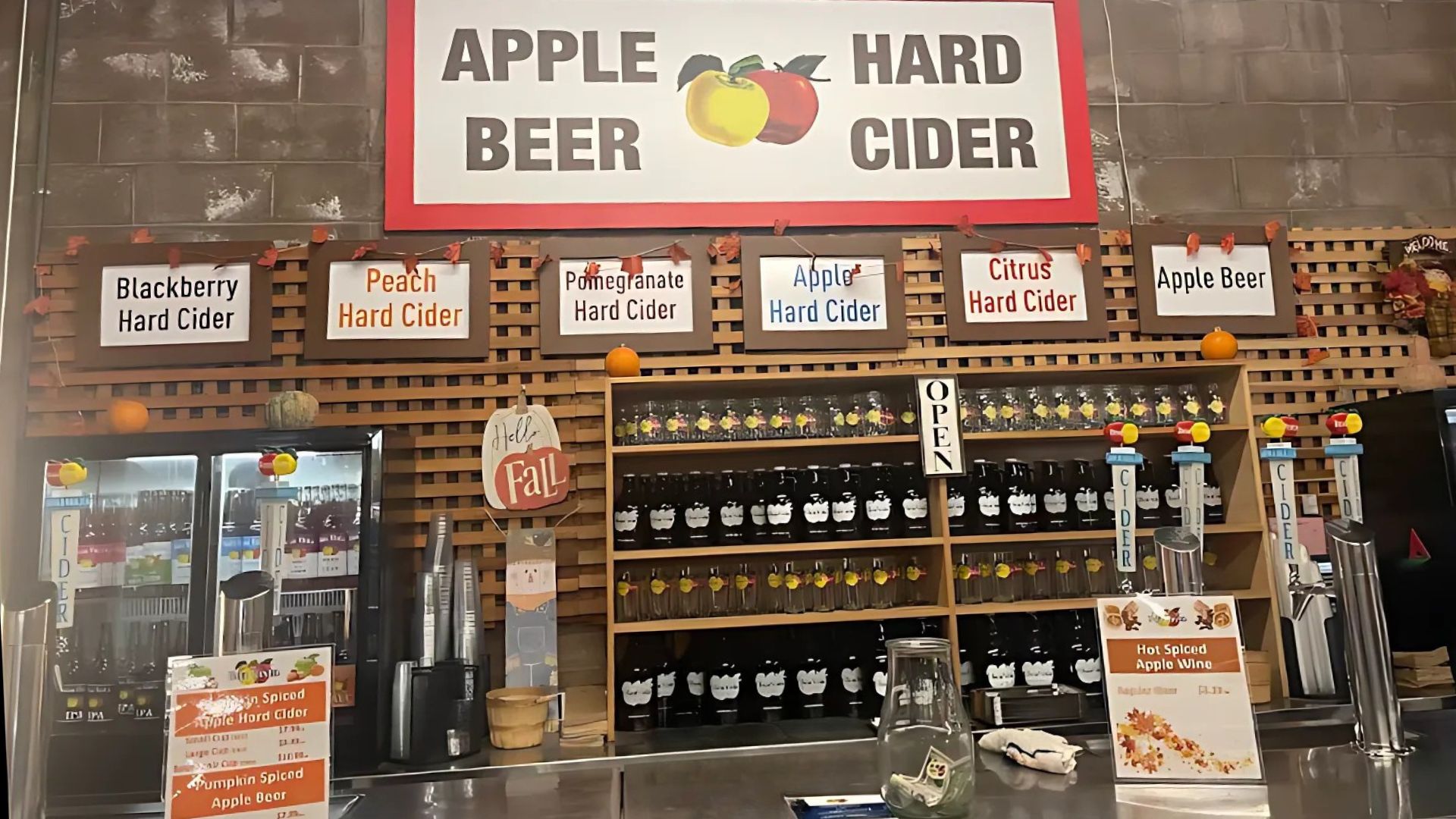 An indoor market stall or counter featuring various apple-based beverages, including hard ciders (blackberry, peach, pomegranate, apple, citrus), apple beer, and hot spiced apple wine. Signs above the counter prominently display "APPLE BEER HARD CIDER" and "OPEN," while shelves behind the counter are stocked with bottles and glasses. Fall-themed decorations, including small pumpkins and autumn leaves, adorn the top of the display. Two refrigerators with more beverages are visible on the left.
