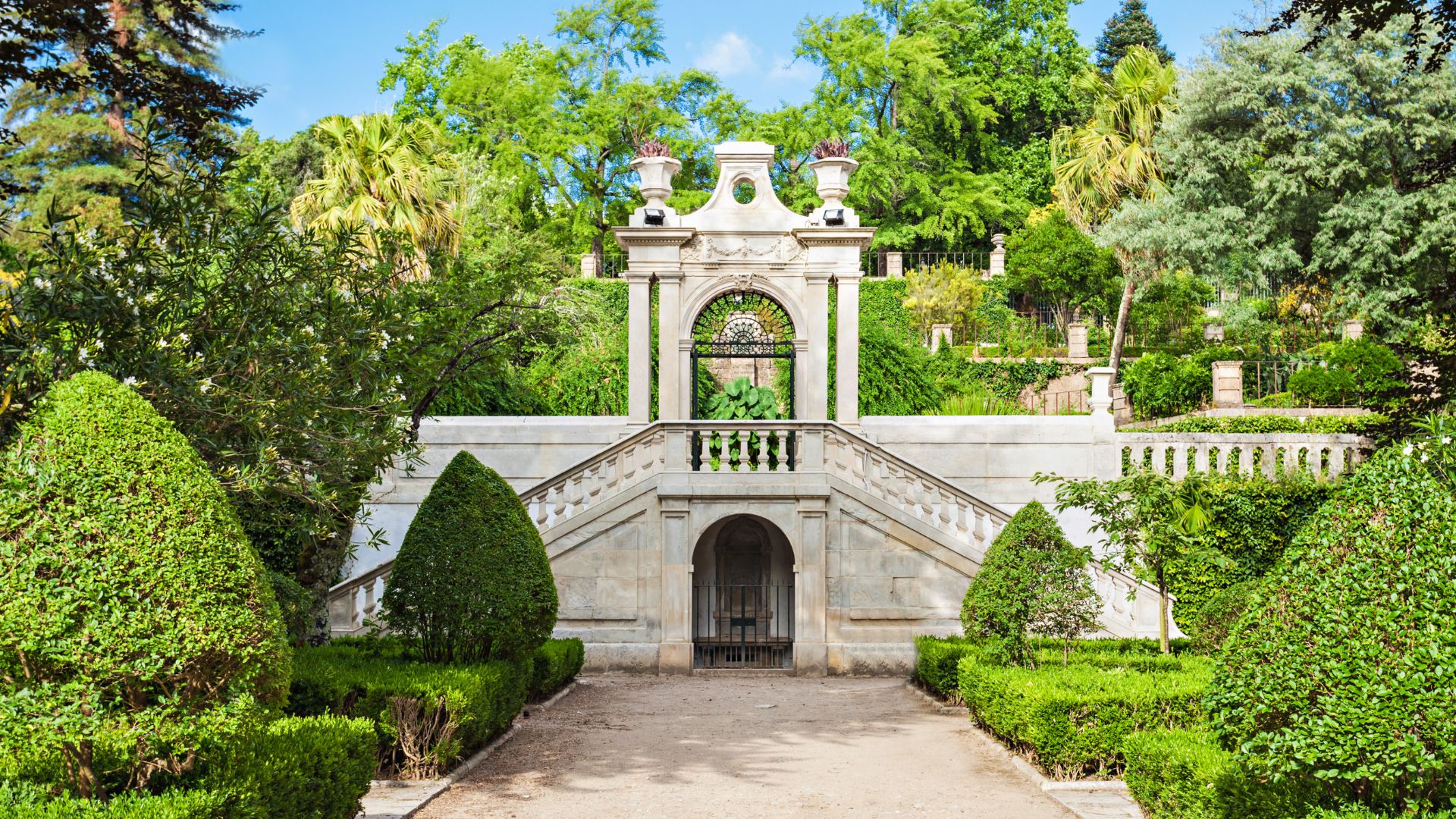 The image showcases a grand stone staircase and entrance gate within the lush and manicured grounds of the Botanical Garden of the University of Coimbra, a historic site in Central Portugal known for its diverse plant collections. 
