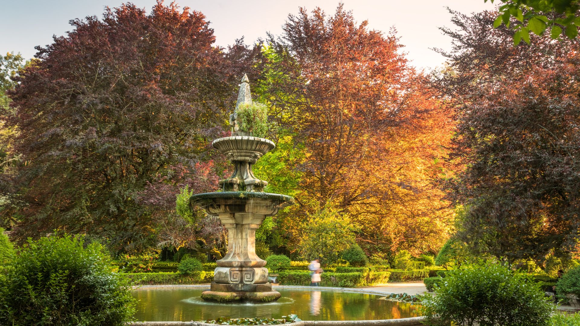 The image captures a serene scene within the Botanical Garden of the University of Coimbra, featuring a multi-tiered stone fountain as its centerpiece, surrounded by lush greenery and vibrant trees, some with reddish foliage, suggesting a beautiful and well-maintained historical garden space.