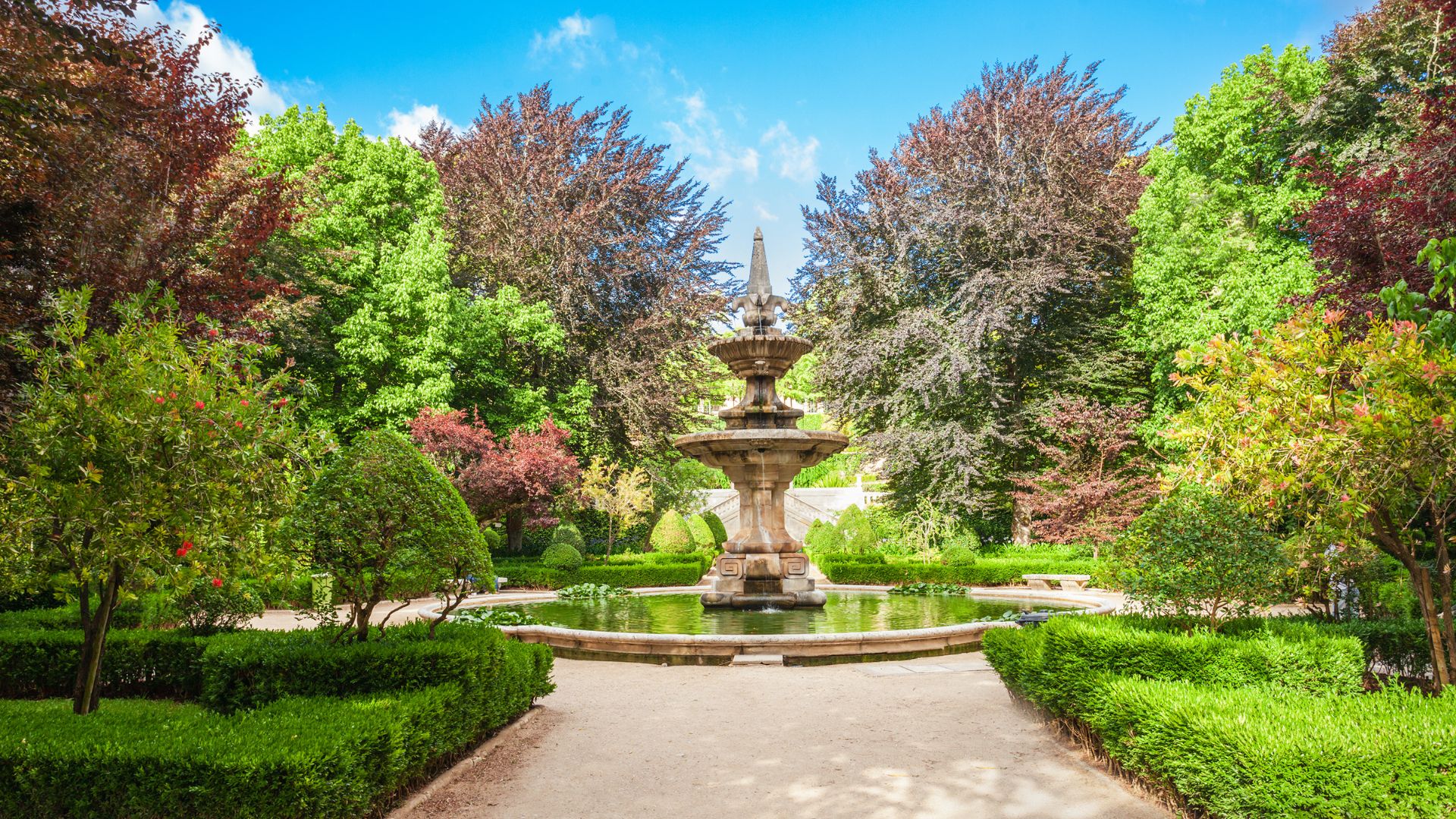 The image captures a serene scene within the Botanical Garden of the University of Coimbra, featuring a multi-tiered stone fountain as its centerpiece, surrounded by lush greenery and vibrant trees, some with reddish foliage, suggesting a beautiful and well-maintained historical garden space.