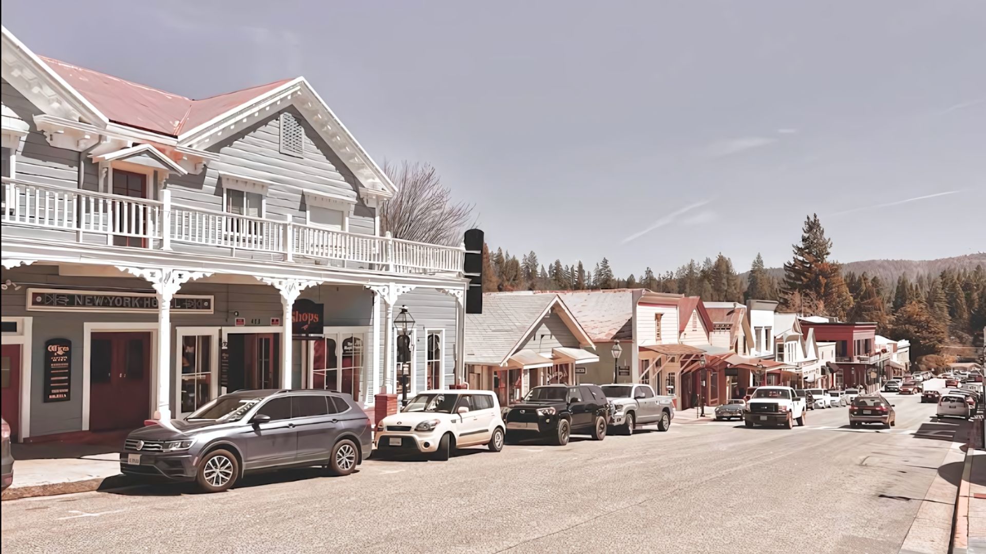 A daytime, eye-level shot of a historic street in a small town, featuring a row of well-preserved 19th-century buildings with intricate details and balconies lining the right side of the road. Several cars are parked along the street, and a few are driving in the distance. Lush green trees and a clear sky with scattered clouds are visible in the background, with hills rising behind the town.