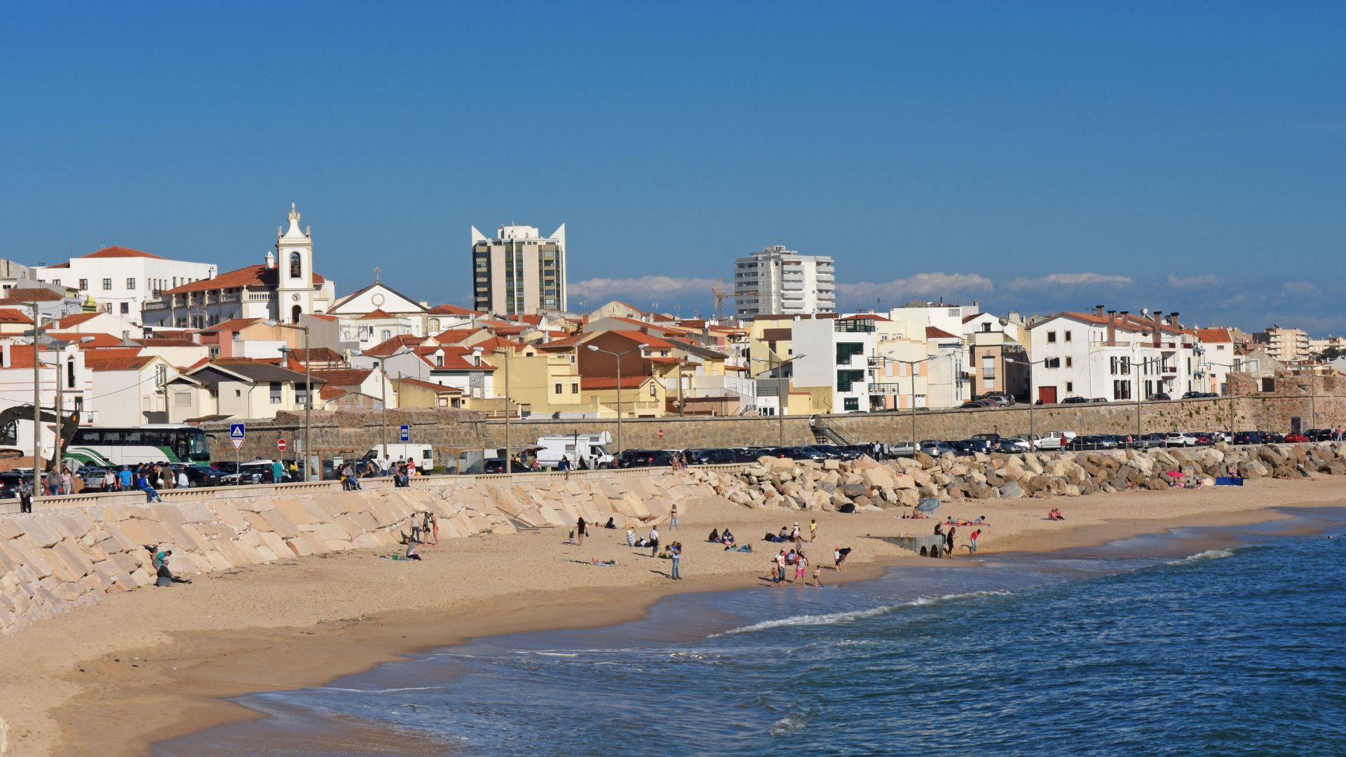 A panoramic view of Buarcos Beach in Figueira da Foz, Portugal, showing a sandy beach with gentle waves, people relaxing on the sand, and a town with buildings and a church along the coastline under a clear blue sky. 