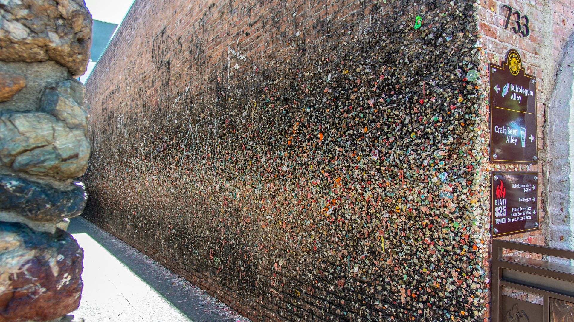 A narrow alleyway with brick walls completely covered in thousands of colorful, chewed pieces of bubblegum, leading towards a brighter opening at the far end. 
