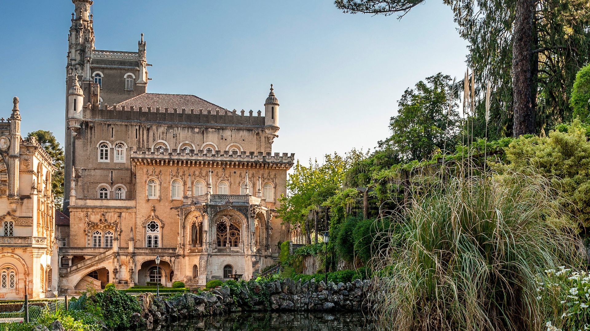 A grand, ornate palace with intricate architectural details, set amidst lush green gardens and trees, with a small pond or water feature in the foreground.