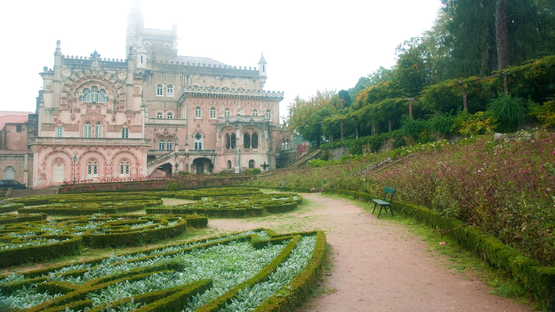 A grand, ornate palace with a light pink facade and intricate architectural details, surrounded by manicured gardens with patterned hedges and a winding dirt path leading towards a lush, forested hillside. The sky is overcast, and a lone bench is visible along the path.