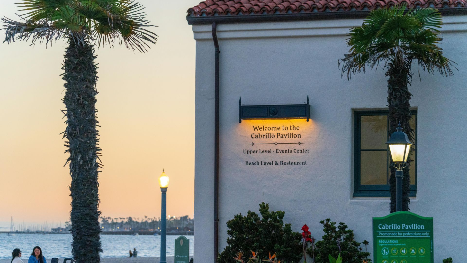 The image captures the exterior of the Cabrillo Pavilion, a historic beachfront venue in Santa Barbara, California, against a warm sunset sky. The building features Spanish Colonial Revival architecture, and a prominent sign welcomes visitors to the "Cabrillo Pavilion" and indicates its "Upper Level - Events Center" and "Beach Level & Restaurant." Palm trees frame the building, and a glimpse of the ocean and beach is visible in the background, along with a streetlamp and a smaller sign for "Cabrillo Pavilion".