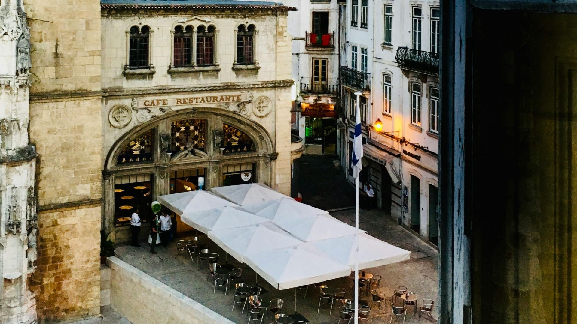 View of the historic city of Coimbra, Portugal, featuring the Monastery of the Holy Cross on the left and Café Santa Cruz with outdoor seating in the foreground, with traditional Portuguese buildings climbing a hill in the background.