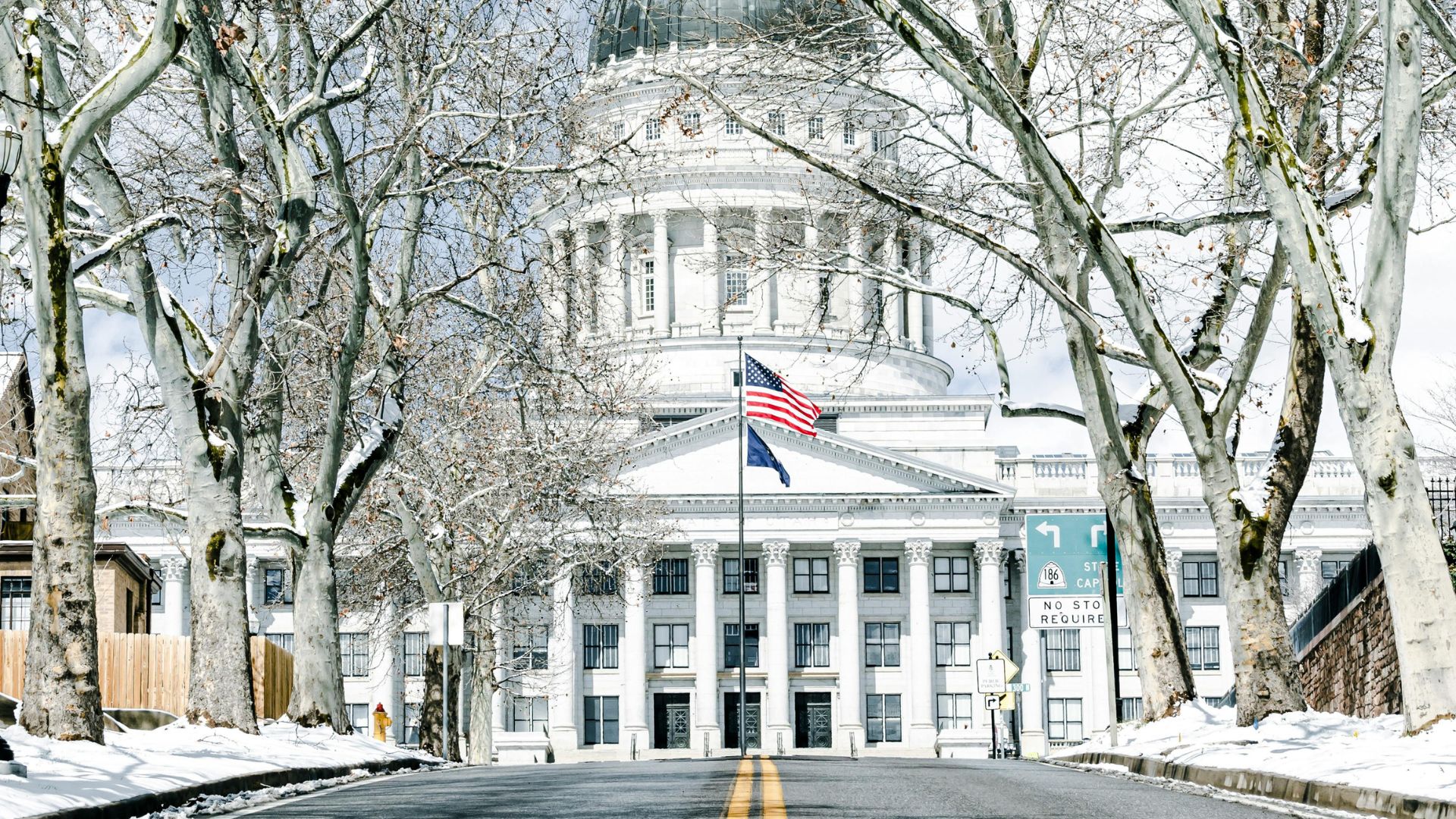 A long, snow-dusted road with a double yellow line leads directly to the grand, white California State Capitol building, which features a large dome and a flag flying in front. Bare, snow-covered trees line both sides of the road, framing the building under a bright, cloudy sky.
