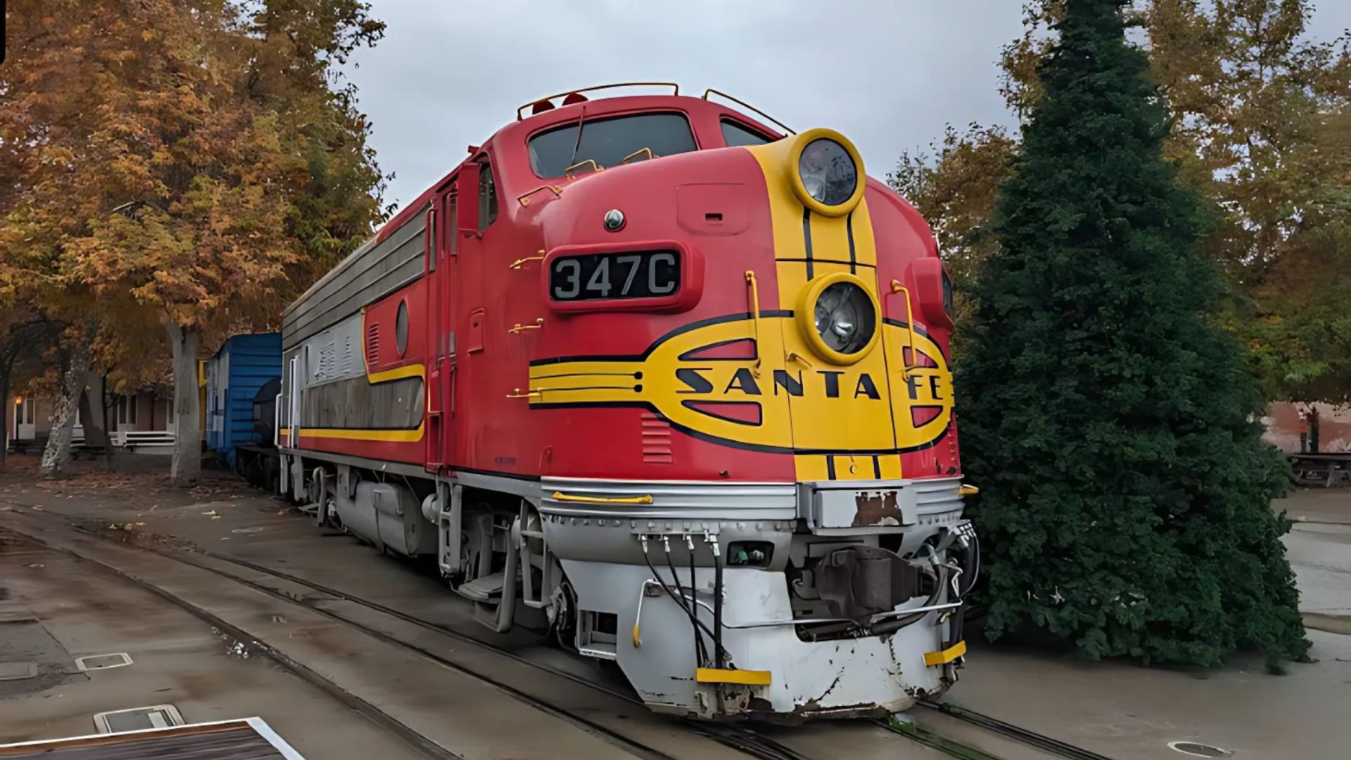 A red and yellow Santa Fe F7 diesel locomotive, identified as 347C, is prominently displayed on tracks outdoors, surrounded by autumn trees and a tall evergreen, under an overcast sky at the California State Railroad Museum in Sacramento.
