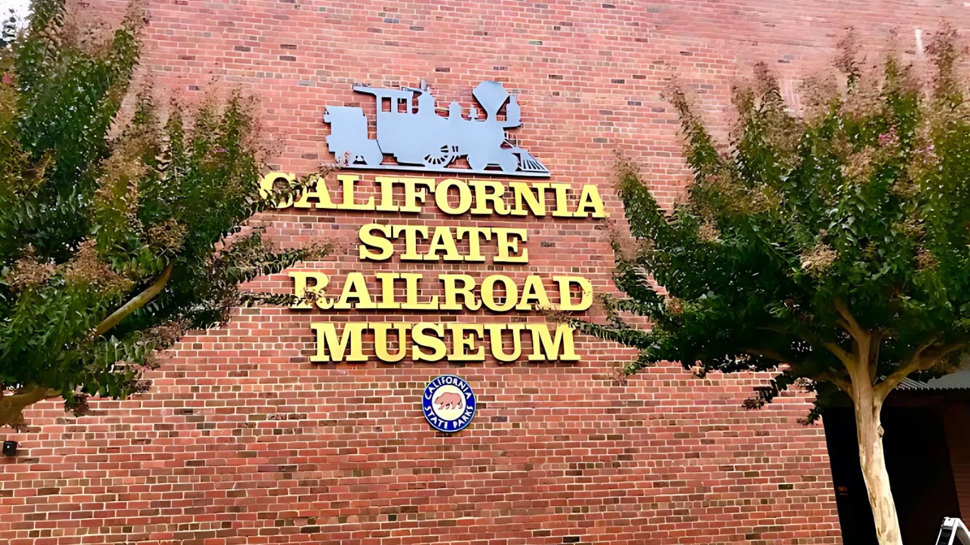 The front exterior of the California State Railroad Museum, featuring a red brick building facade with large golden letters spelling "CALIFORNIA STATE RAILROAD MUSEUM" and a blue train silhouette above the text. A round blue and white emblem is centered below the text, and two green trees flank the building entrance.