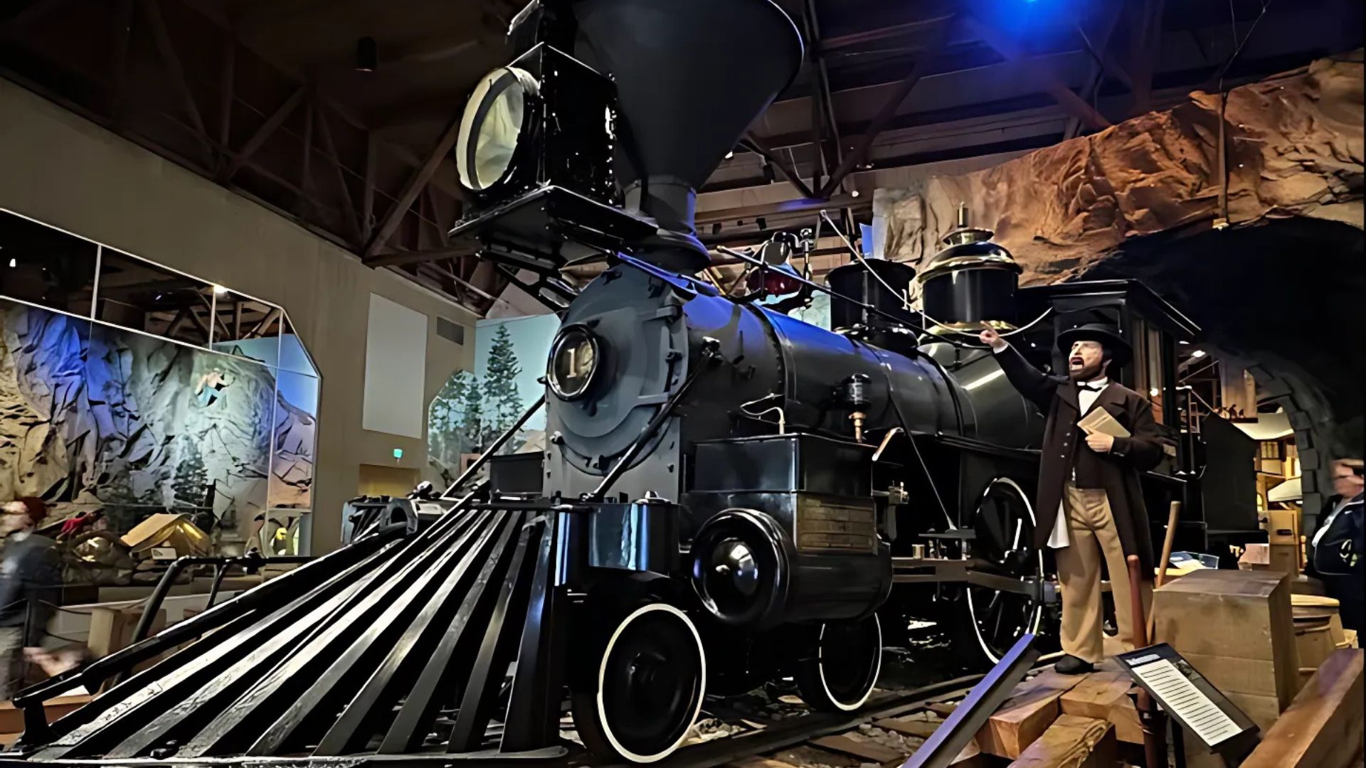 A large, black vintage steam locomotive with a prominent funnel and cowcatcher is displayed inside a museum, with a mannequin in historical clothing standing near its front right side. The background shows exhibits depicting natural landscapes and possibly a railroad station setting under blue lighting.