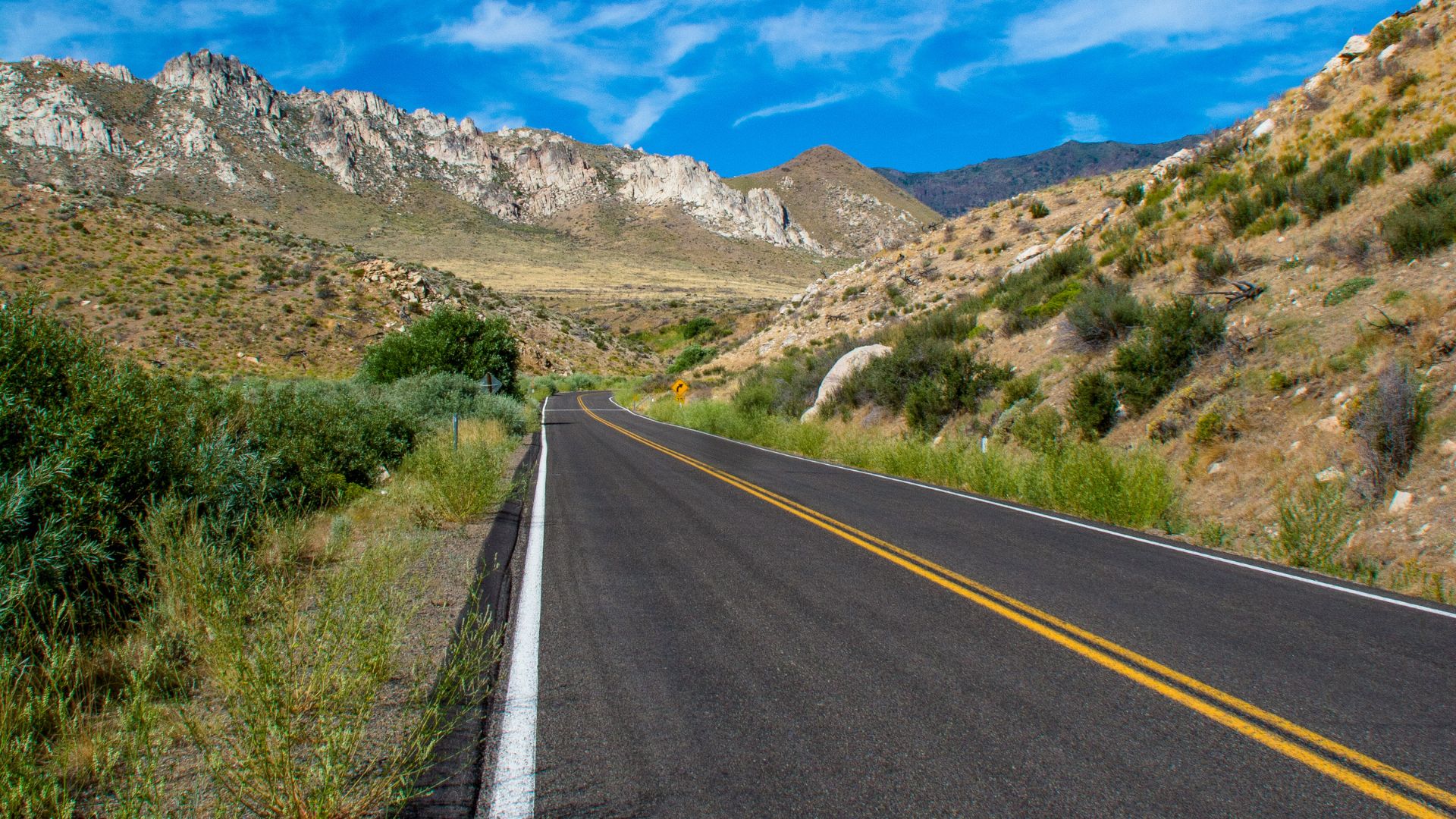 A paved road with double yellow lines and a white shoulder line extends through a mountainous, arid landscape under a clear blue sky.