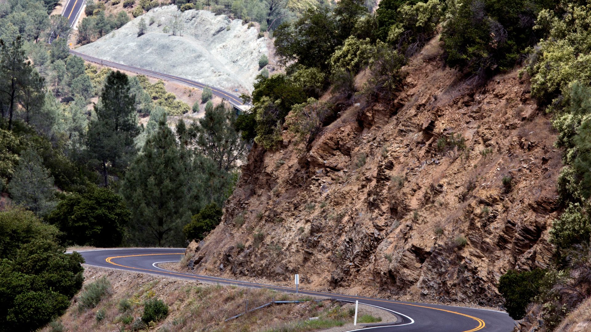 A winding, two-lane road with a yellow center line curves sharply through a mountainous landscape covered in trees and rocky terrain under a bright sky. Another section of the road is visible in the distance on a lighter-colored hillside.