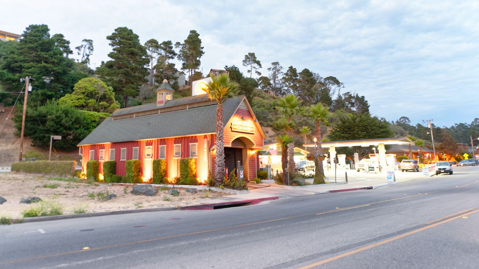 A brightly lit, barn-like building with a red exterior and a dark roof, illuminated by numerous lights, stands next to a road and a gas station at dusk in Cambria, California. Palm trees and other greenery are visible around the building and on the hillside in the background.