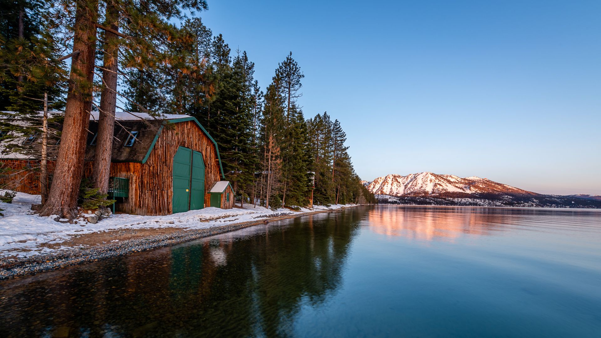 A rustic wooden boathouse with green doors stands on the snowy shore of Lake Tahoe, with a snow-capped mountain reflecting in the calm water under a clear sky at dawn.