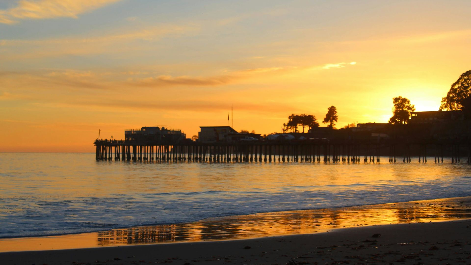 A vibrant sunset casts warm orange and yellow hues across the sky and reflects on the calm water of Capitola Beach, with the silhouette of Capitola Wharf and structures extending into the ocean from the right, against a backdrop of dark land and silhouetted trees.
