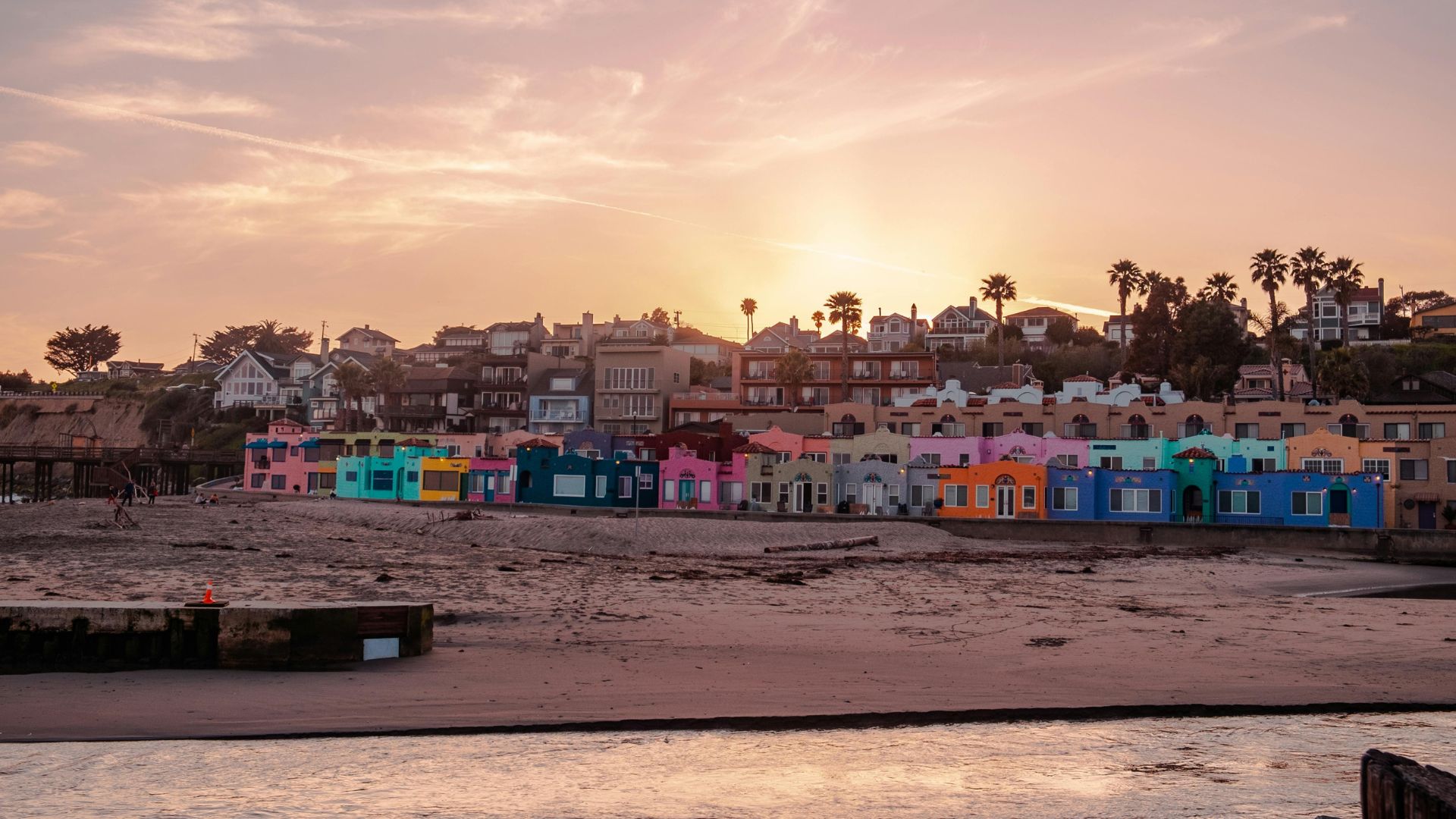 A panoramic view of Capitola Beach at sunset, featuring a row of vibrant, colorful houses along the sandy shore with palm trees and other buildings on a hillside in the background, under a warm, cloudy sky.