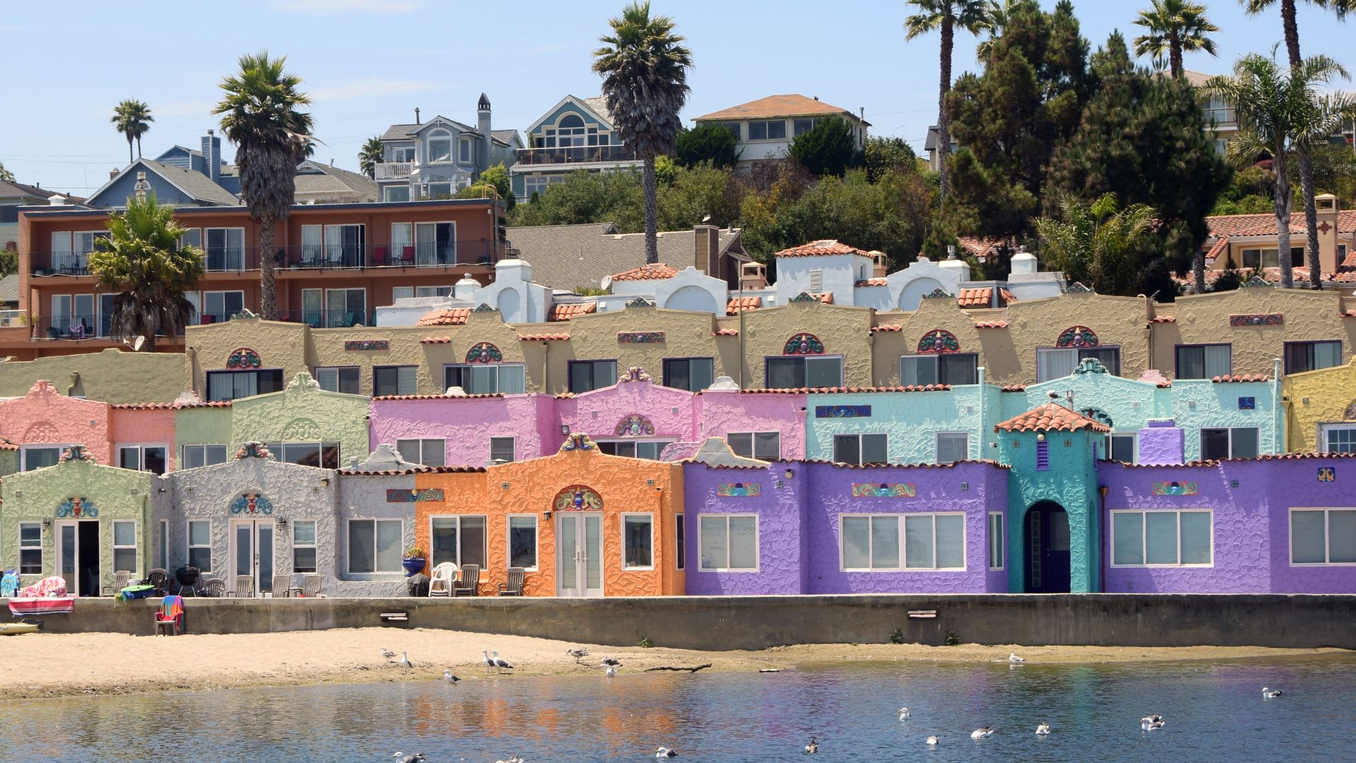An eye-level shot captures the colorful Venetian Court condominiums and the Capitola Venetian Hotel reflecting in the calm waters of the Capitola Lagoon at sunset. The pastel-colored buildings, ranging from pinks and oranges to blues and greens, line the waterfront, with palm trees and additional houses visible on the hillside above under a dramatic sky with hues of orange and purple. 