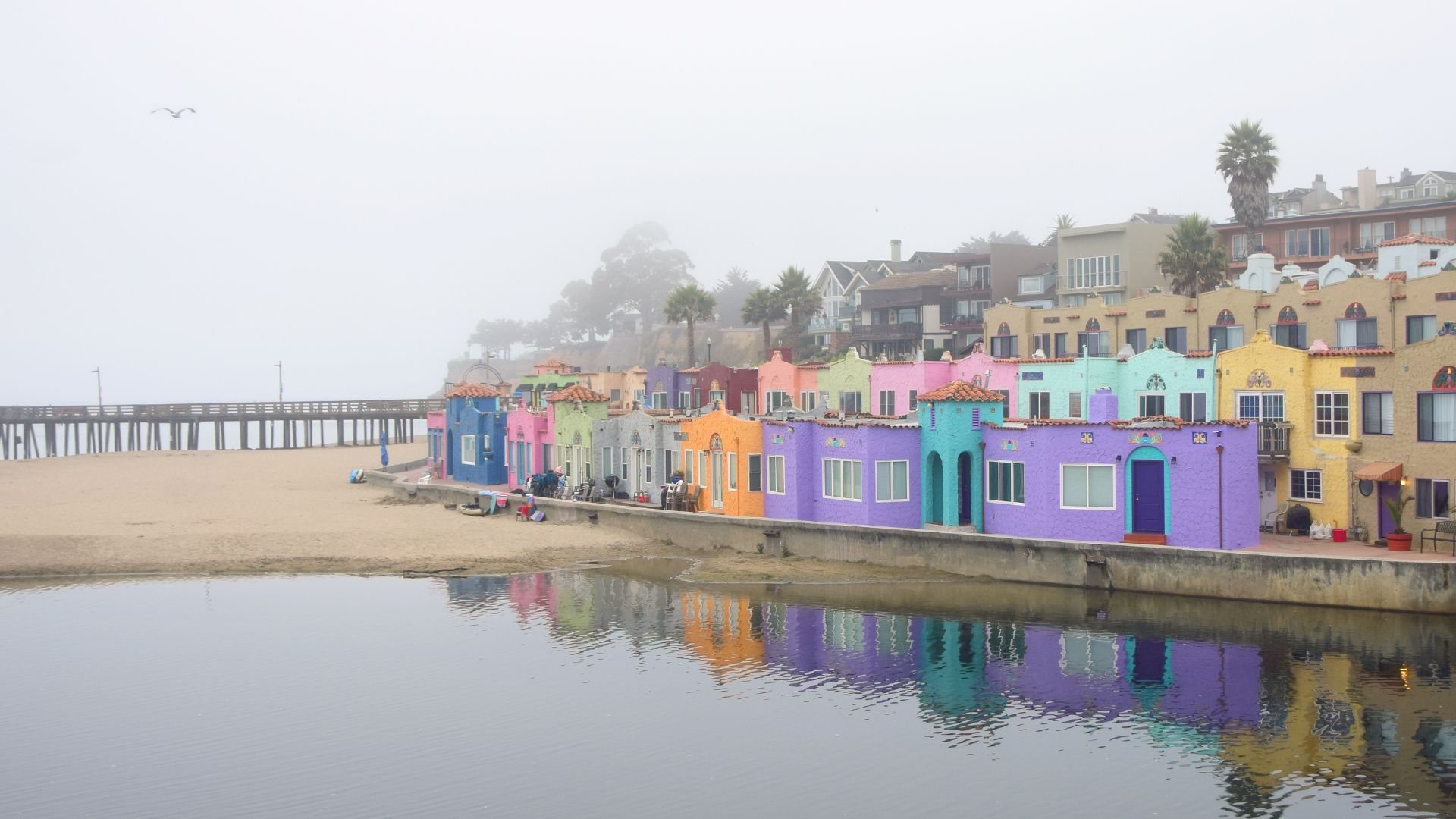 A panoramic view of the colorful Venetian Court buildings in Capitola, California, lining a sandy beach and calm body of water, likely the Capitola Lagoon or Monterey Bay, under a hazy or foggy sky. A long pier extends into the distance on the left, and reflections of the vibrant buildings are visible in the water.
