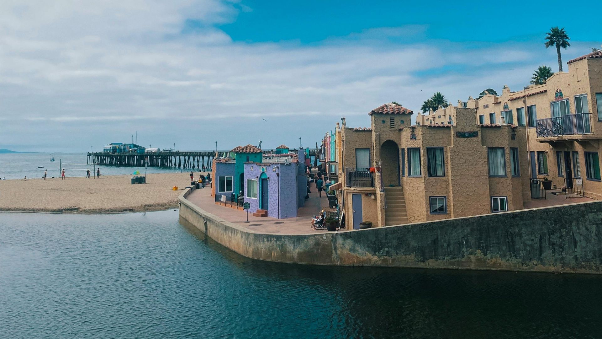 A vibrant daytime view of Capitola Beach in California, featuring the colorful Venetian Court apartments lining a calm waterway that opens to the Pacific Ocean. In the background, a sandy beach extends towards a pier, under a partially cloudy blue sky.