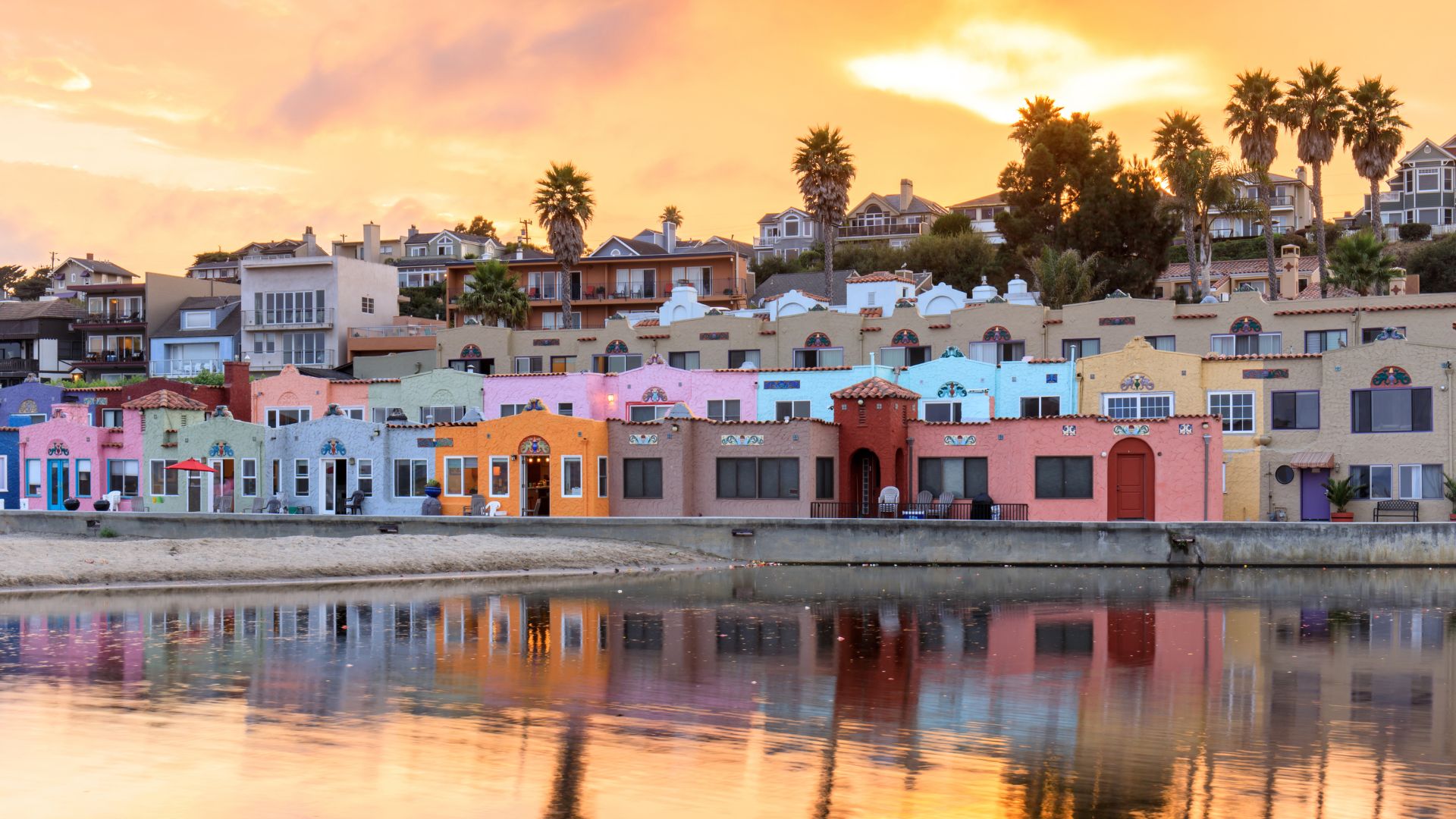 A vibrant sunset illuminates the colorful buildings of Capitola Village along the waterfront, with palm trees and houses on a hill in the background, all reflected in the calm water in the foreground. 