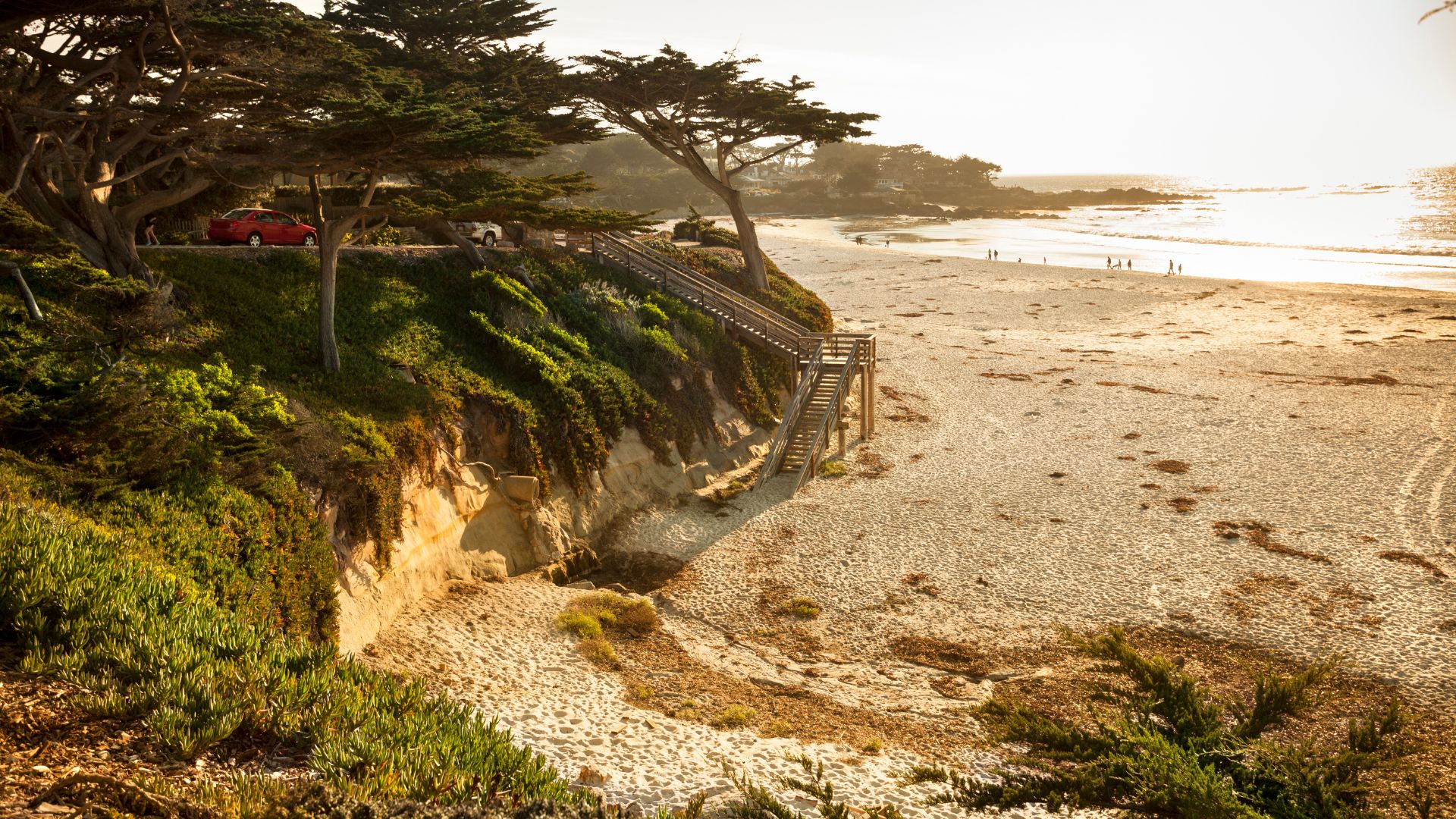 A sunlit sandy beach with gentle waves meeting the shore, bordered by a steep, green-covered cliff on the left, featuring a wooden staircase leading down to the sand and cypress trees at the top of the cliff. A red car is visible on a road at the top of the cliff.