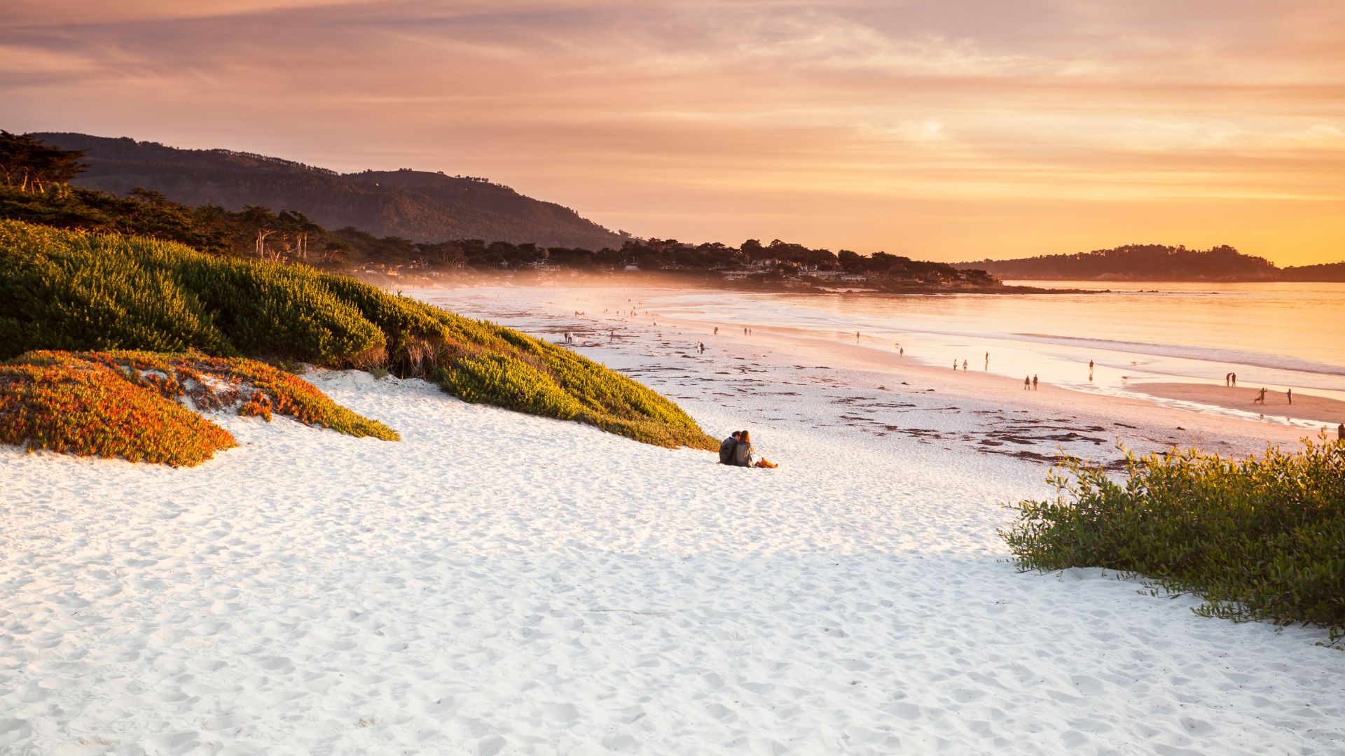 A wide shot of a white-sand beach with calm ocean waters under a vibrant sunset sky, featuring a person sitting on the sand and lush green and orange-tinged vegetation on the dune to the left.