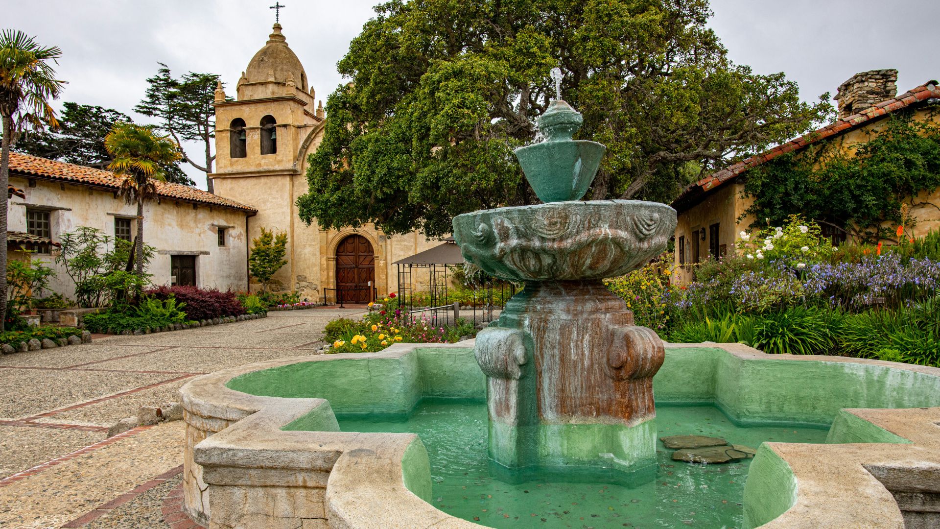 A historic Spanish mission featuring a stone bell tower, a courtyard with a large tiered fountain, and lush green landscaping under a cloudy sky. The architecture includes adobe-style buildings and a prominent church structure.