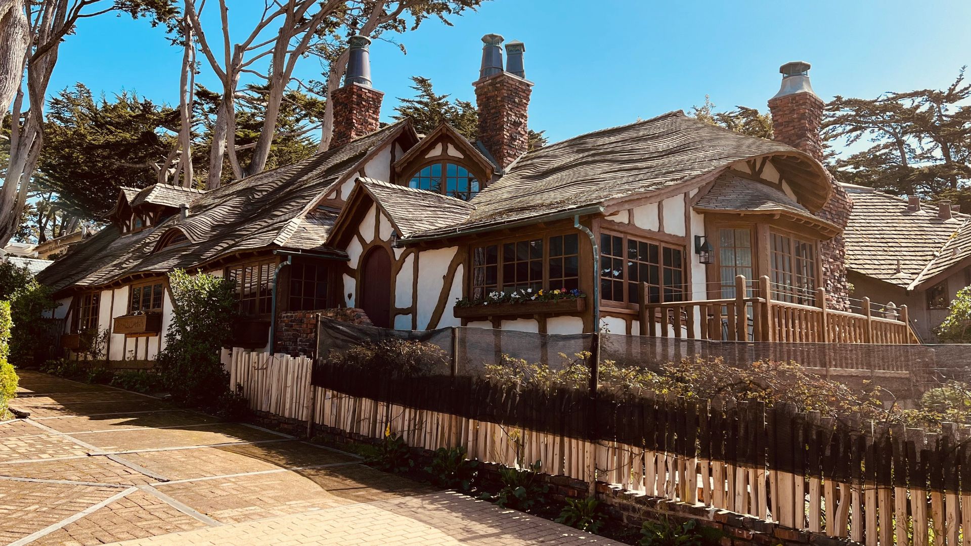 A charming, multi-level storybook-style house with a distinctive wavy roof, stone chimneys, and wooden accents, nestled among trees on a sunny day in Carmel-by-the-Sea, California.