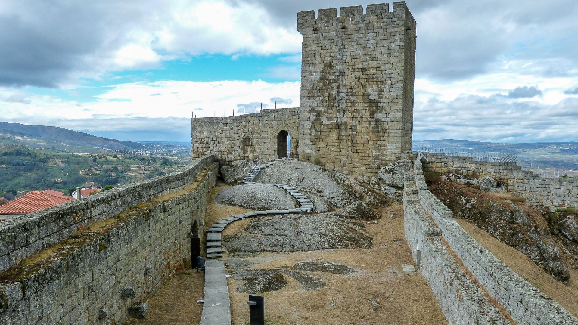 The image depicts the medieval Castle of Linhares da Beira, a historic fortress situated on high ground, showcasing its formidable stone walls, a prominent keep (Torre de Menagem), and a pathway leading up to its entrance. 