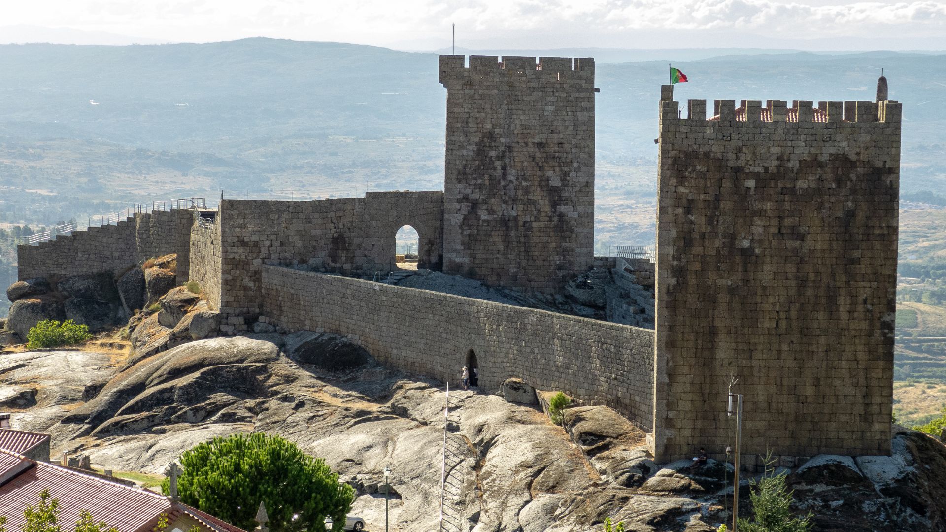 A medieval stone castle with two prominent towers and connecting walls, perched on a rocky outcrop overlooking a valley.