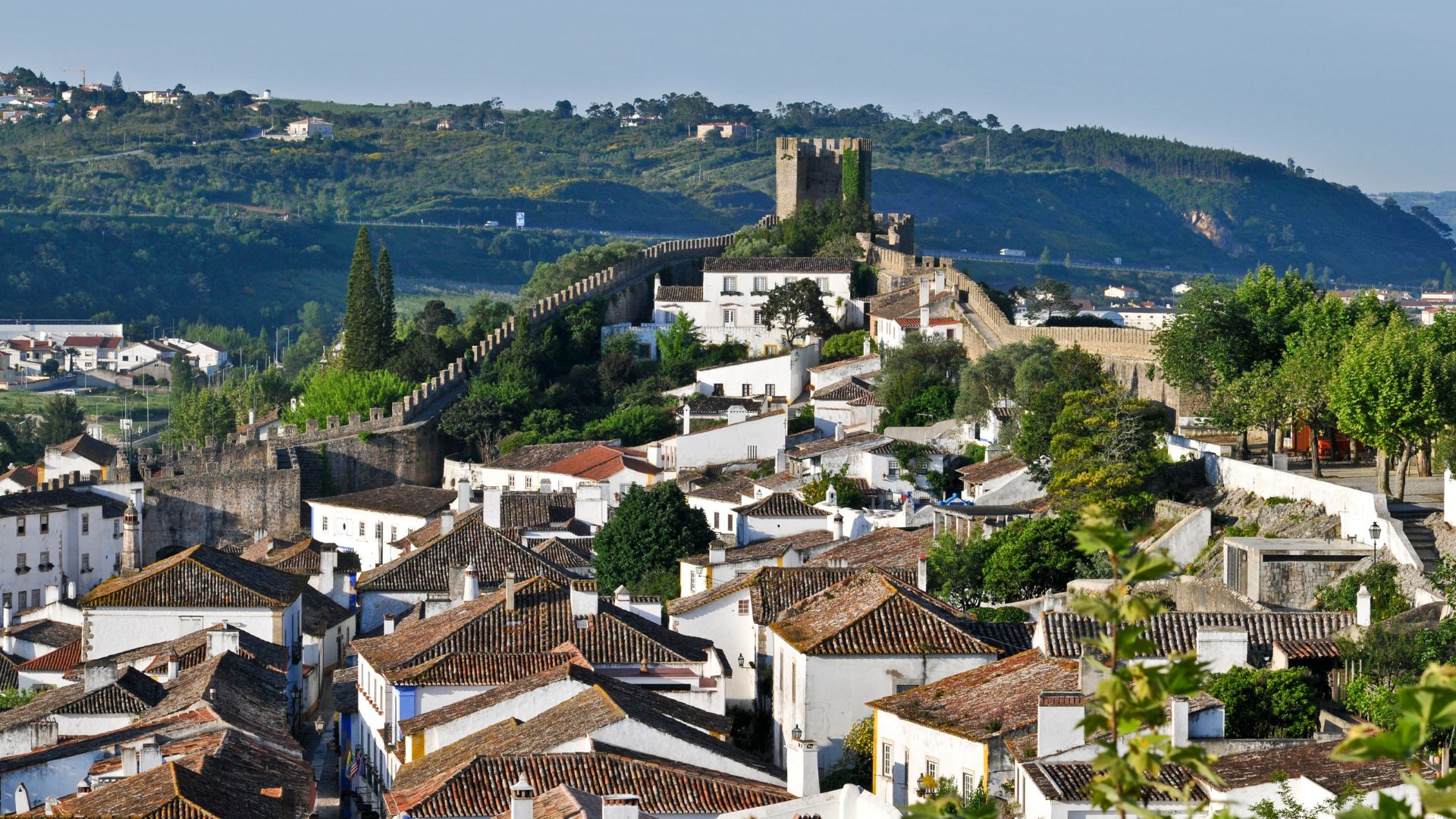 A panoramic view of the medieval walled town of Óbidos, Portugal, with the historic Castelo de Óbidos prominently situated on a hilltop, surrounded by whitewashed buildings with terracotta roofs, lush greenery, and a clear blue sky.