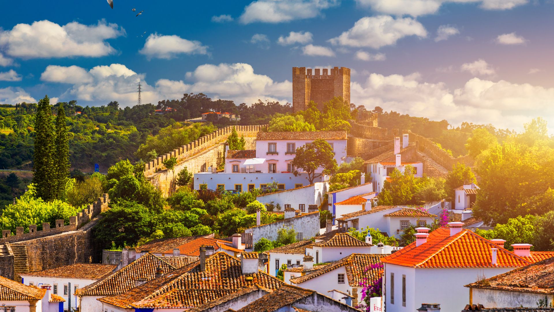 A panoramic view of the medieval walled town of Óbidos, Portugal, with the historic Castelo de Óbidos prominently situated on a hilltop, surrounded by whitewashed buildings with terracotta roofs, lush greenery, and a clear blue sky with scattered clouds.