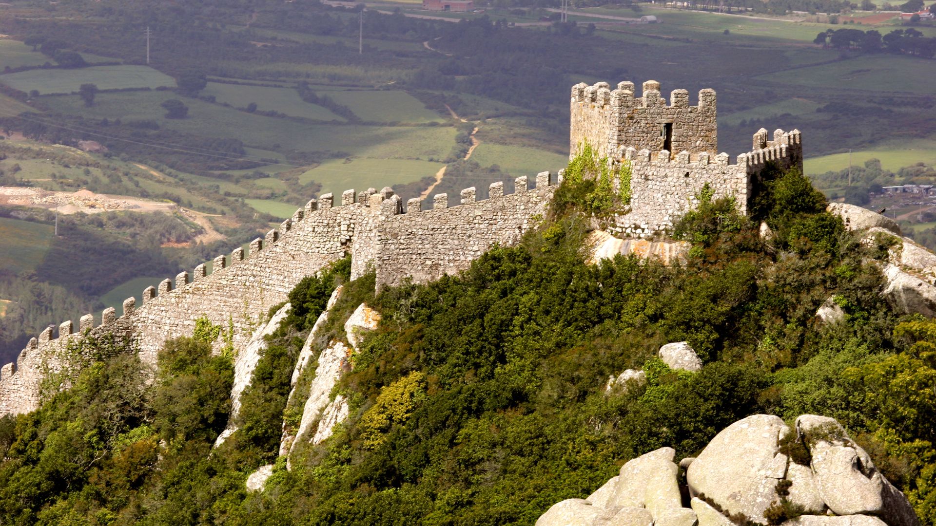 A panoramic view of the ancient Castelo dos Mouros (Moorish Castle) in Sintra, Portugal, showcasing its long stone walls and towers winding along a lush, rocky hilltop with rolling green hills in the background.