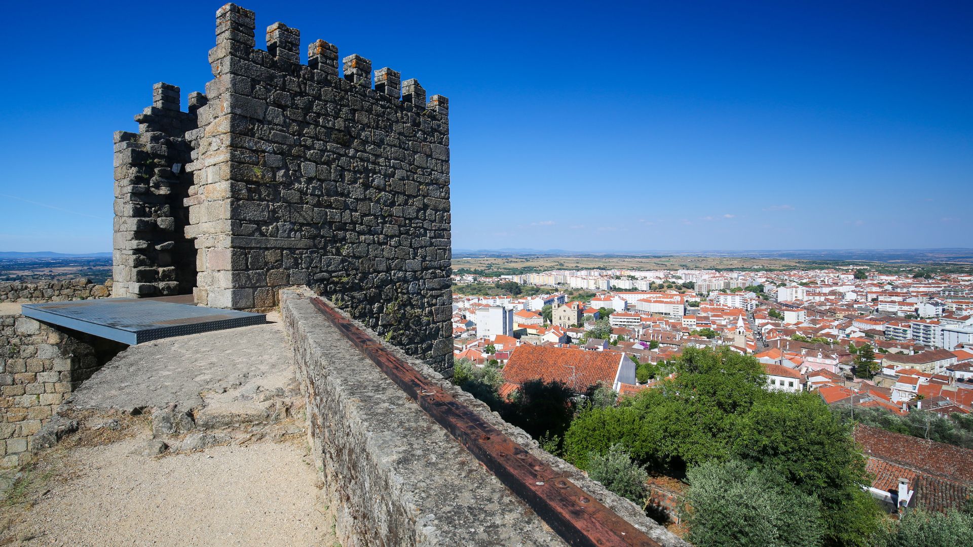 A stone castle fortification with a crenellated wall in the foreground, overlooking a city with red-tiled roofs and a vast landscape under a clear blue sky.