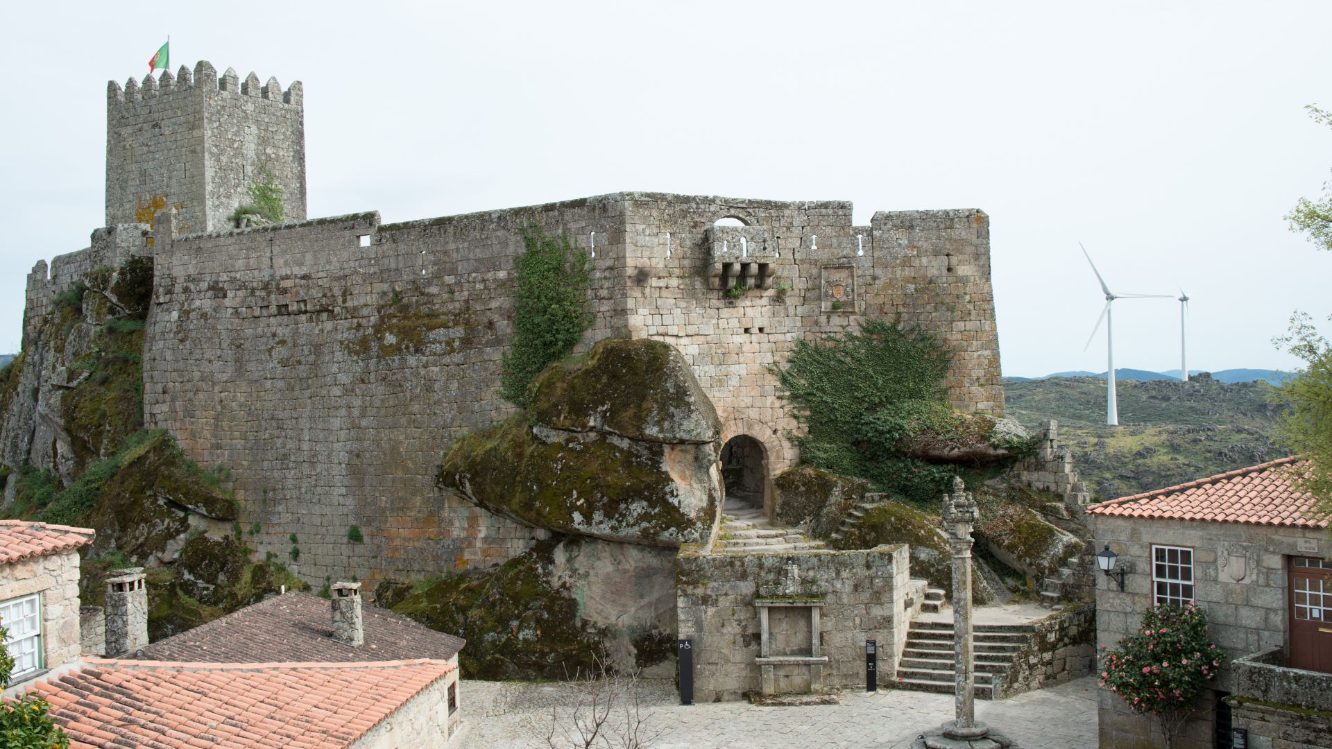 A wide shot of the medieval Castle of Sortelha in Portugal, built on a granite massif with a prominent keep and fortified walls, overlooking a village with traditional stone houses and distant wind turbines.