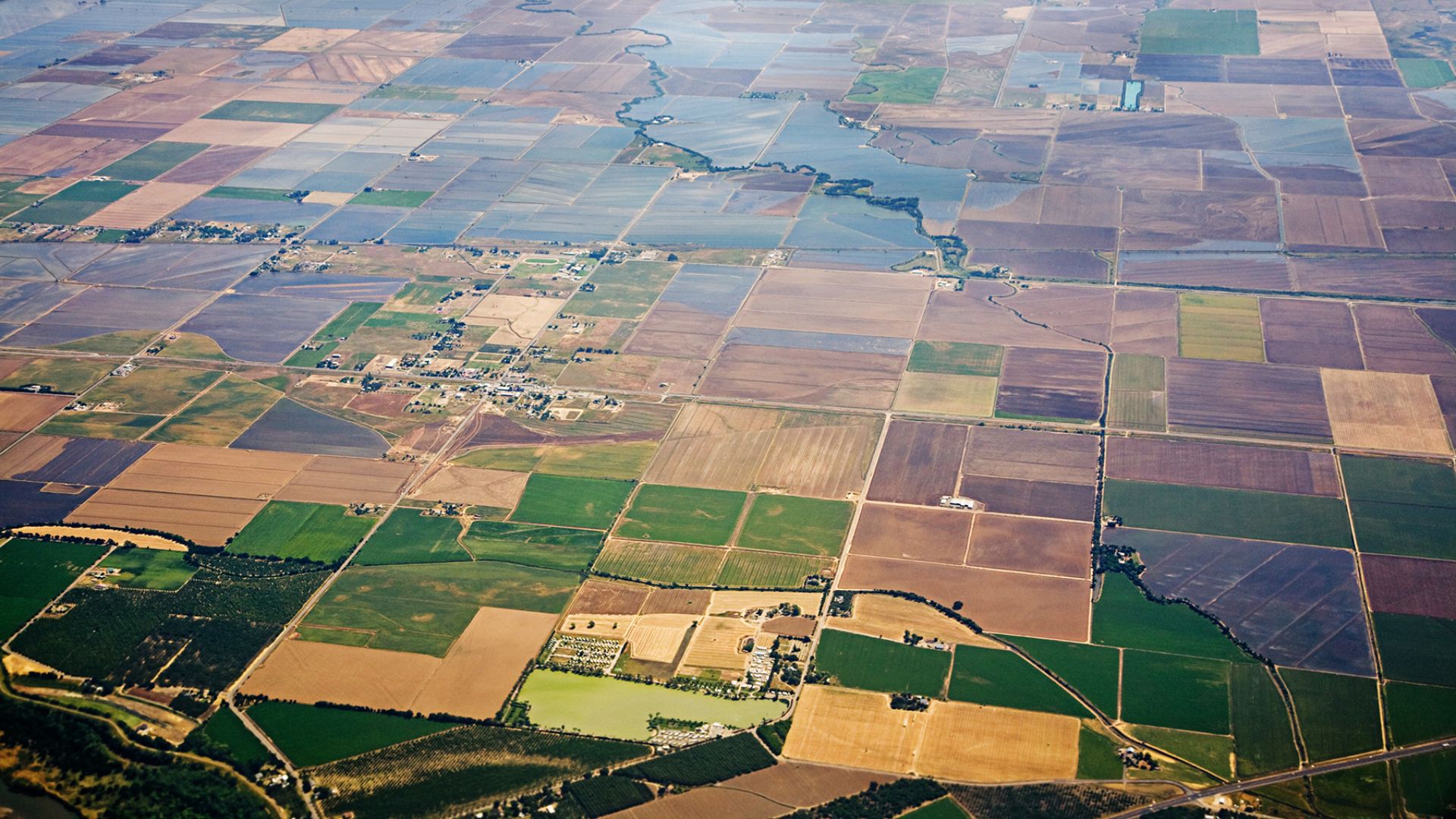 An aerial view of the Central Valley in California, featuring a patchwork of large, rectangular agricultural fields in varying shades of green and brown, interspersed with irrigation canals and a winding river. A small cluster of buildings is visible in the middle ground, indicating a rural community within the agricultural landscape.