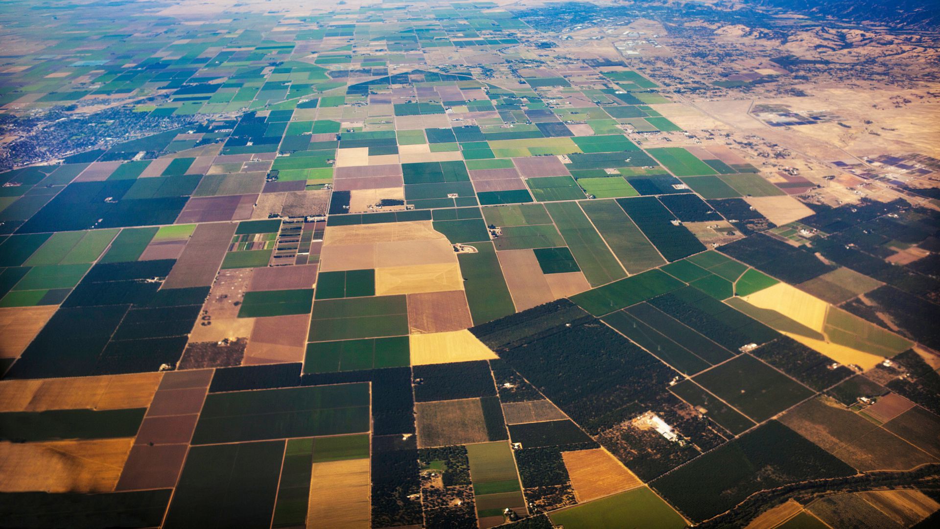 An aerial view of the Central Valley in California, featuring a patchwork of large, rectangular agricultural fields in varying shades of green and brown, interspersed with irrigation canals and a winding river. A small cluster of buildings is visible in the middle ground, indicating a rural community within the agricultural landscape.