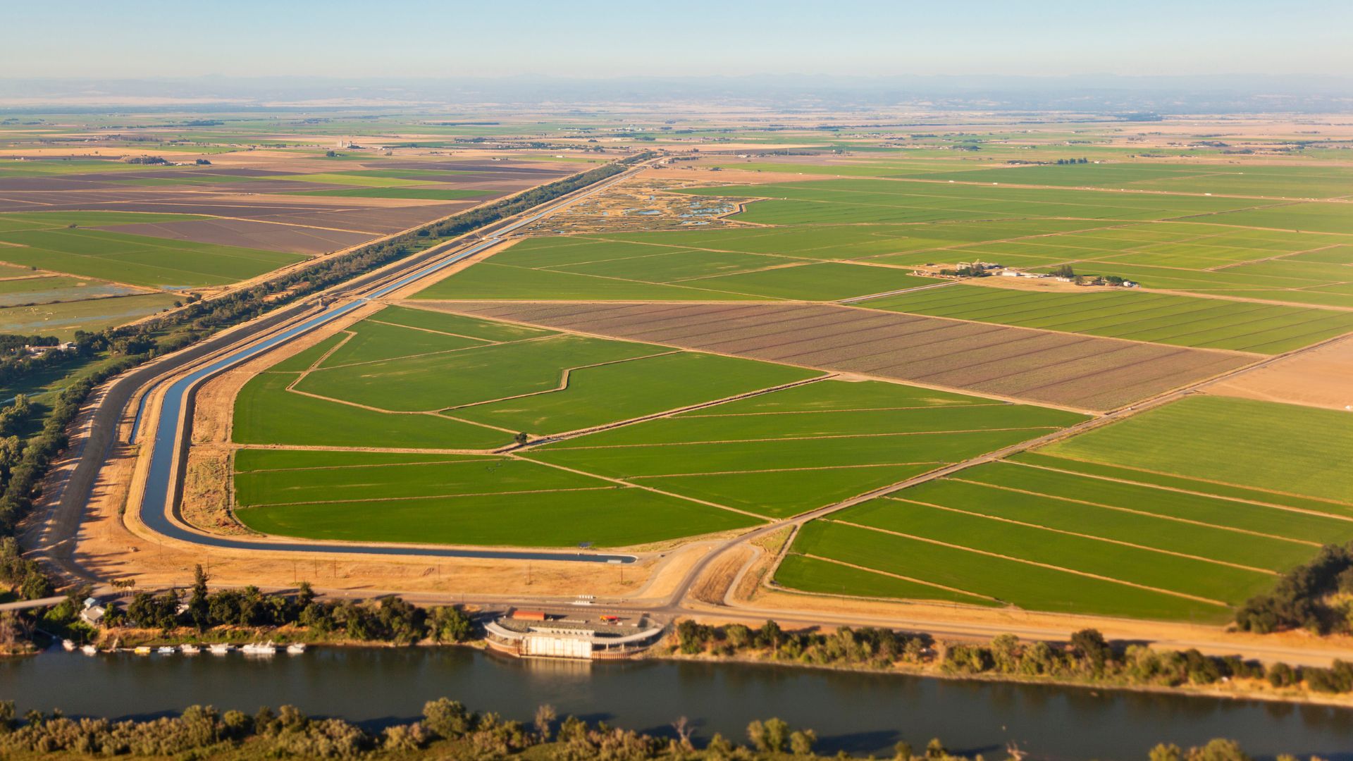 An aerial view of the Central Valley in California shows vast agricultural fields in varying shades of green and brown, indicating different crops and stages of cultivation. A prominent irrigation canal snakes through the landscape, connecting to a wider river in the foreground where a building complex with a dock is visible. A distant mountain range is faintly visible on the horizon.