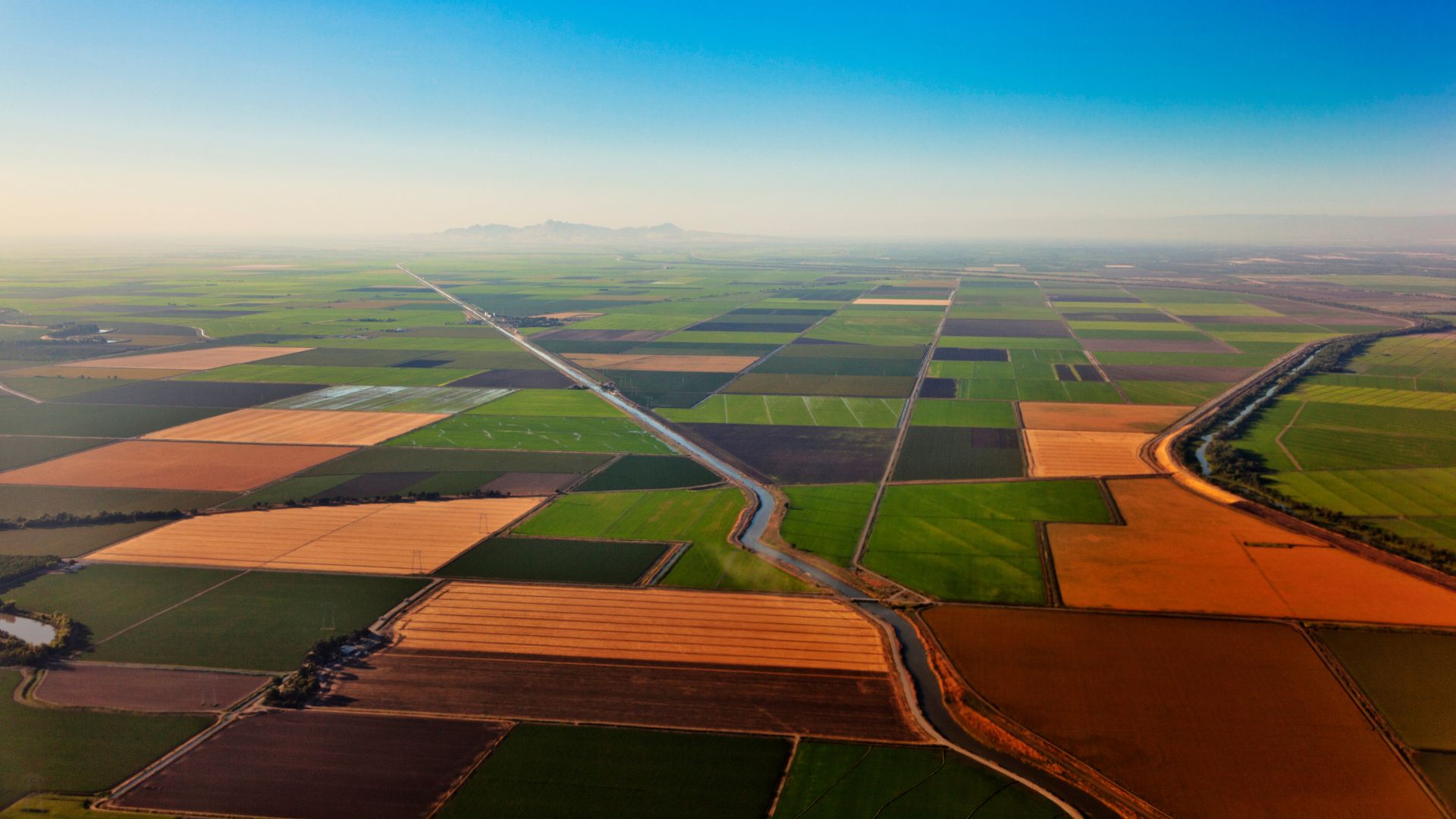 An aerial view of vast agricultural fields in California's Central Valley, featuring a patchwork of green and golden-brown cultivated land, traversed by irrigation canals and a straight road, with distant mountains visible under a clear sky. 