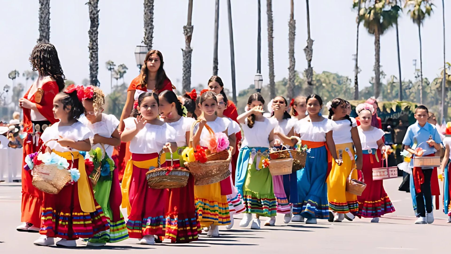 A group of children in colorful traditional Spanish and Mexican attire, carrying baskets, participate in the Children's Parade during the Old Spanish Days Fiesta in Santa Barbara, California, with palm trees lining the background.