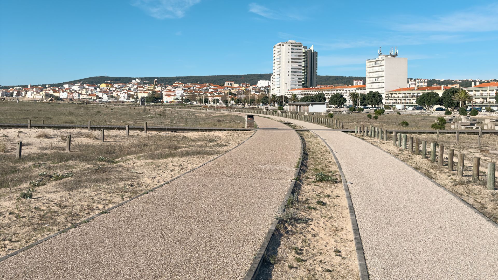 A view of a wide, sandy beach with pedestrian walkways leading towards a cityscape featuring tall buildings under a clear blue sky. In the background, a town sprawls across hills.