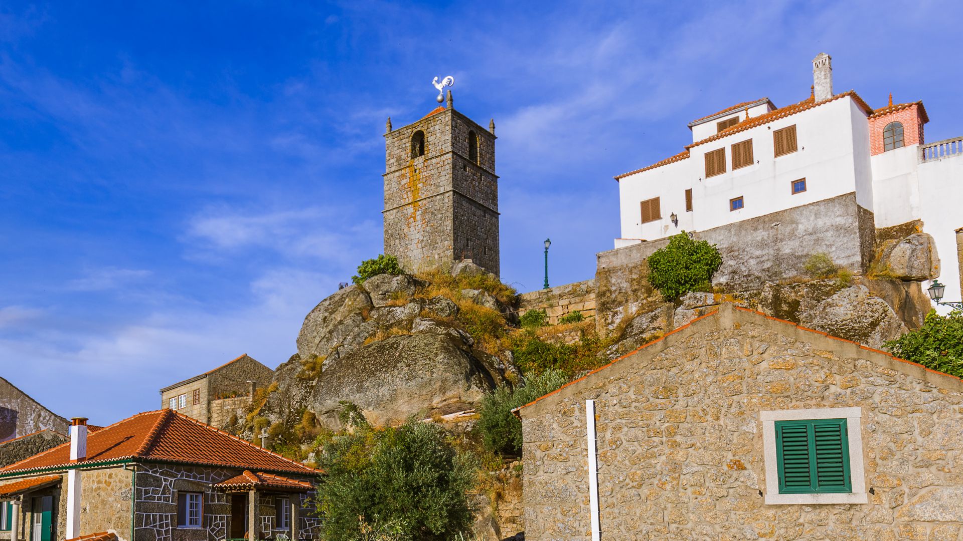A stone clock tower with a white rooster on top, situated on a rocky hill in Monsanto, Portugal, with traditional stone houses and a clear blue sky.
