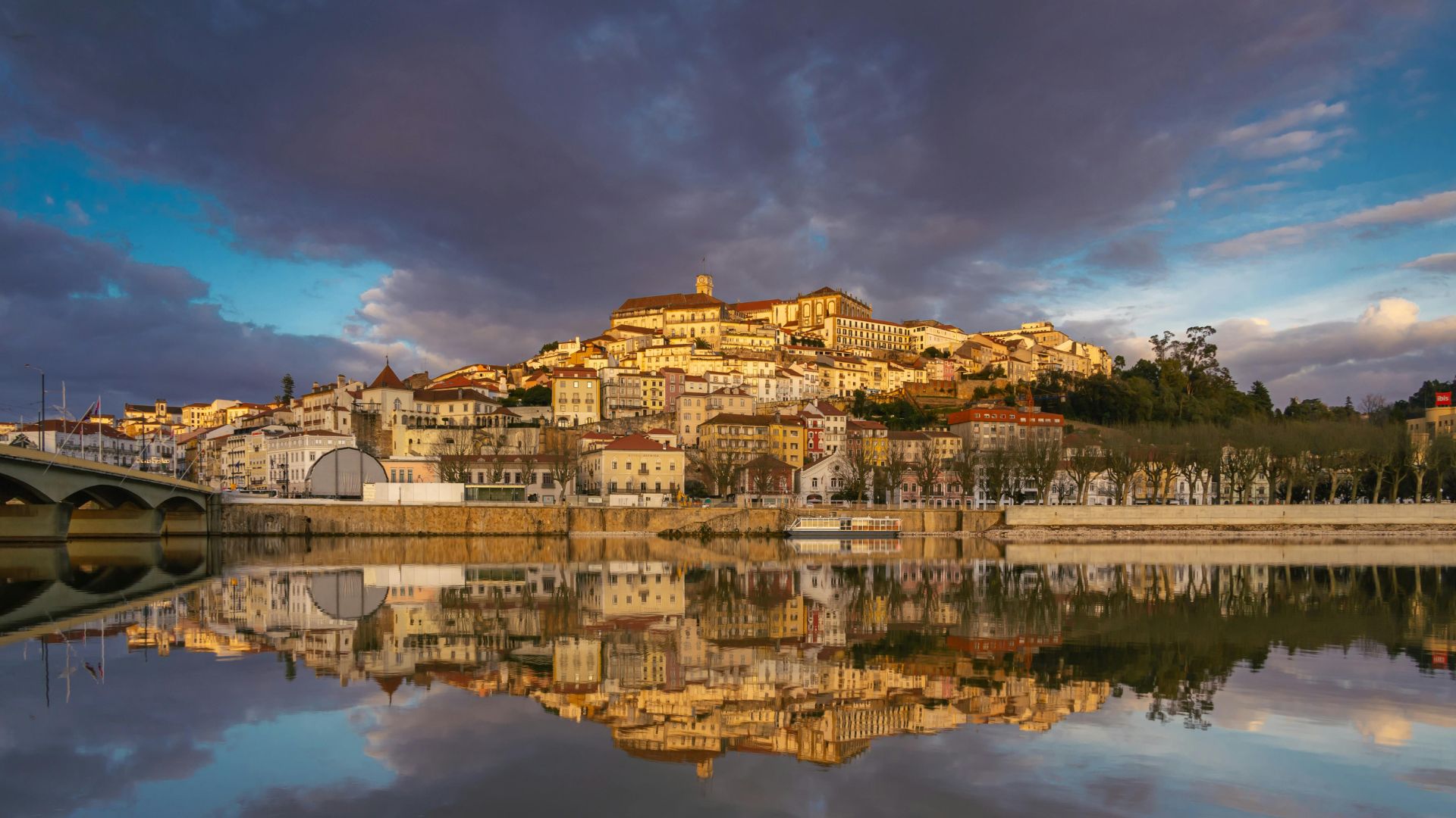 A panoramic view of Coimbra, Portugal, featuring the city's historic buildings reflecting in the calm waters of the Mondego River under a dramatic sky.