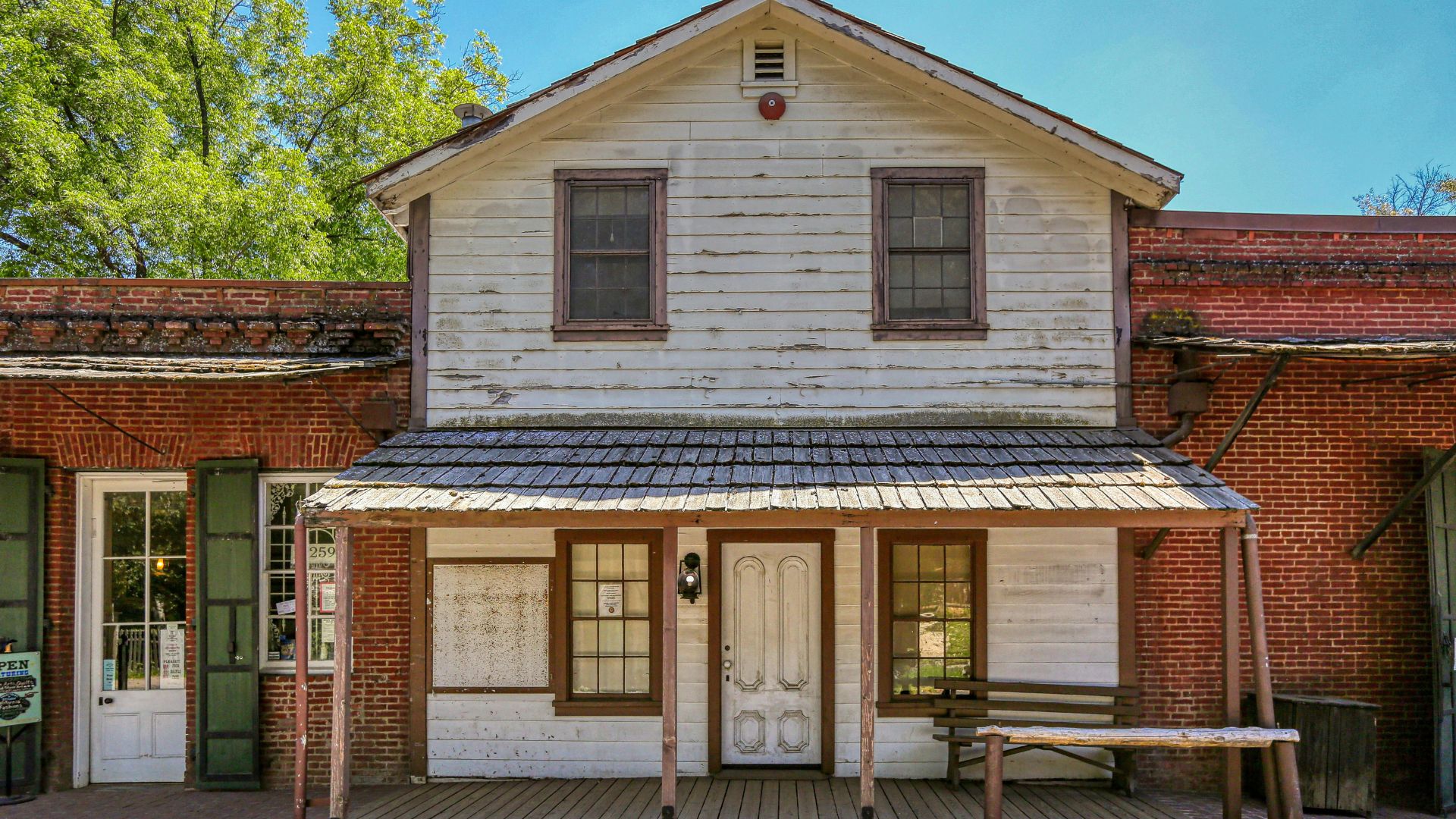 A front-facing view of a historic two-story wooden building with a weathered white facade and a shingled porch roof, featuring multiple windows and a central doorway, flanked by red brick buildings and green trees under a clear sky.