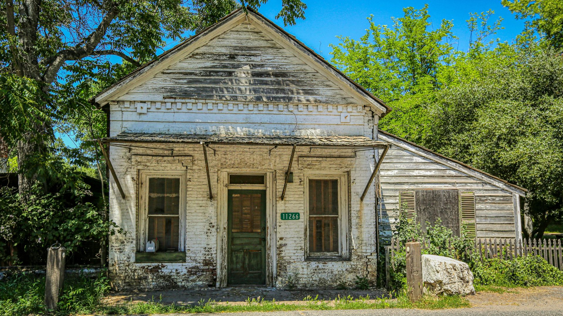 A weathered, white-painted wooden building with a green door and two windows on the front, set against a backdrop of green trees and a clear blue sky. The building appears to be an old commercial structure, possibly a former store or office, with a number "11266" displayed above the door. A smaller, weathered wooden structure is attached to the right side of the main building.